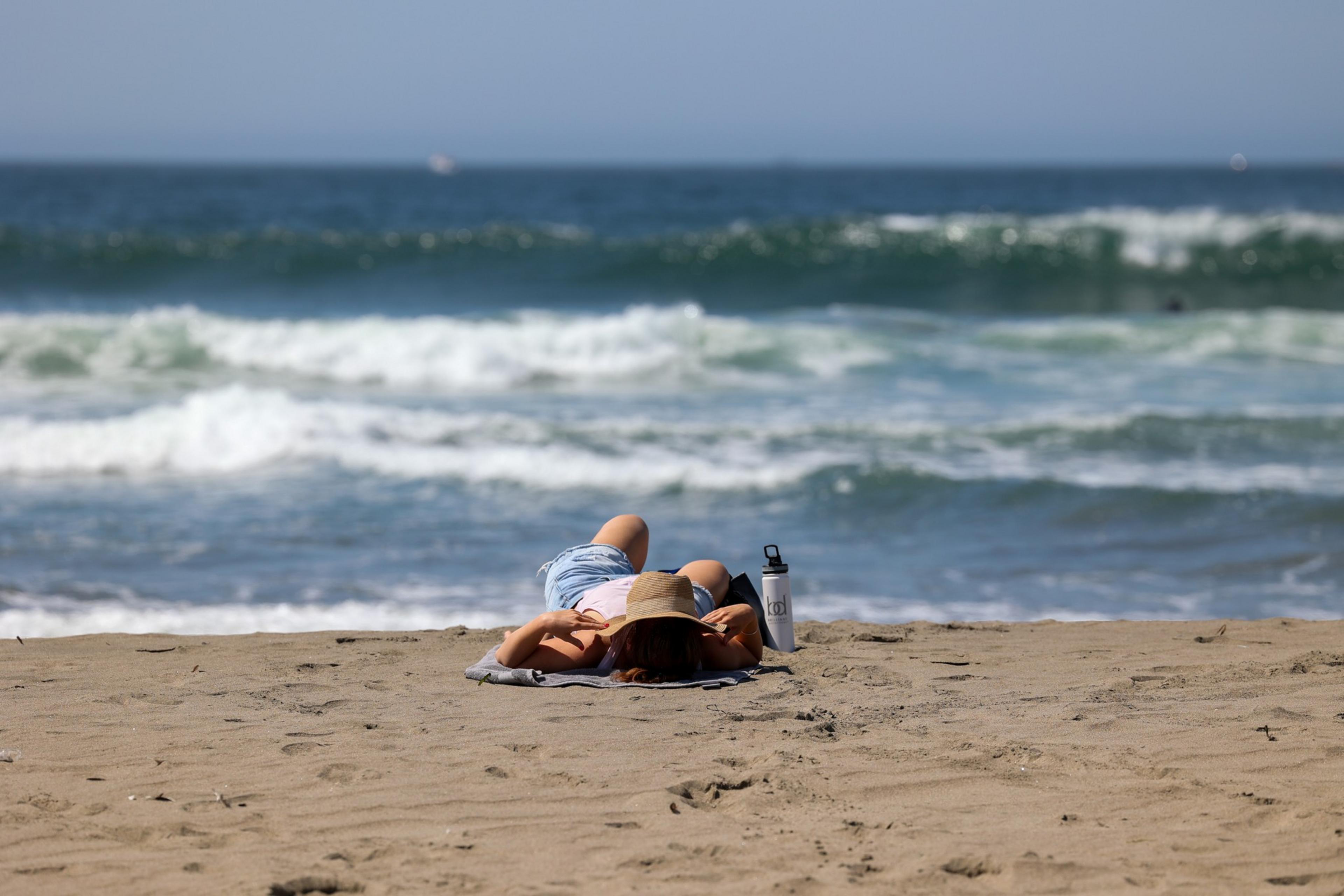 A person lies face down on a towel on the sandy beach, wearing a hat and shorts, with ocean waves breaking in the background.