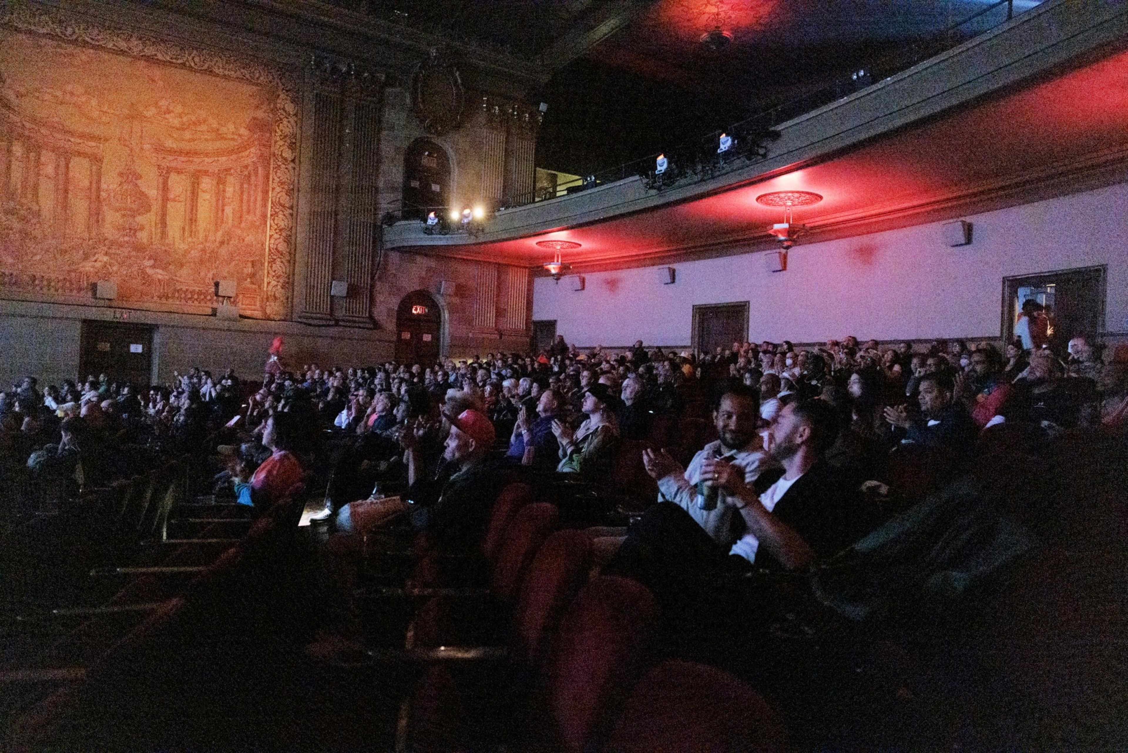A large audience is seated in a dimly lit theater with ornate walls and a balcony, attentively watching a performance or event.