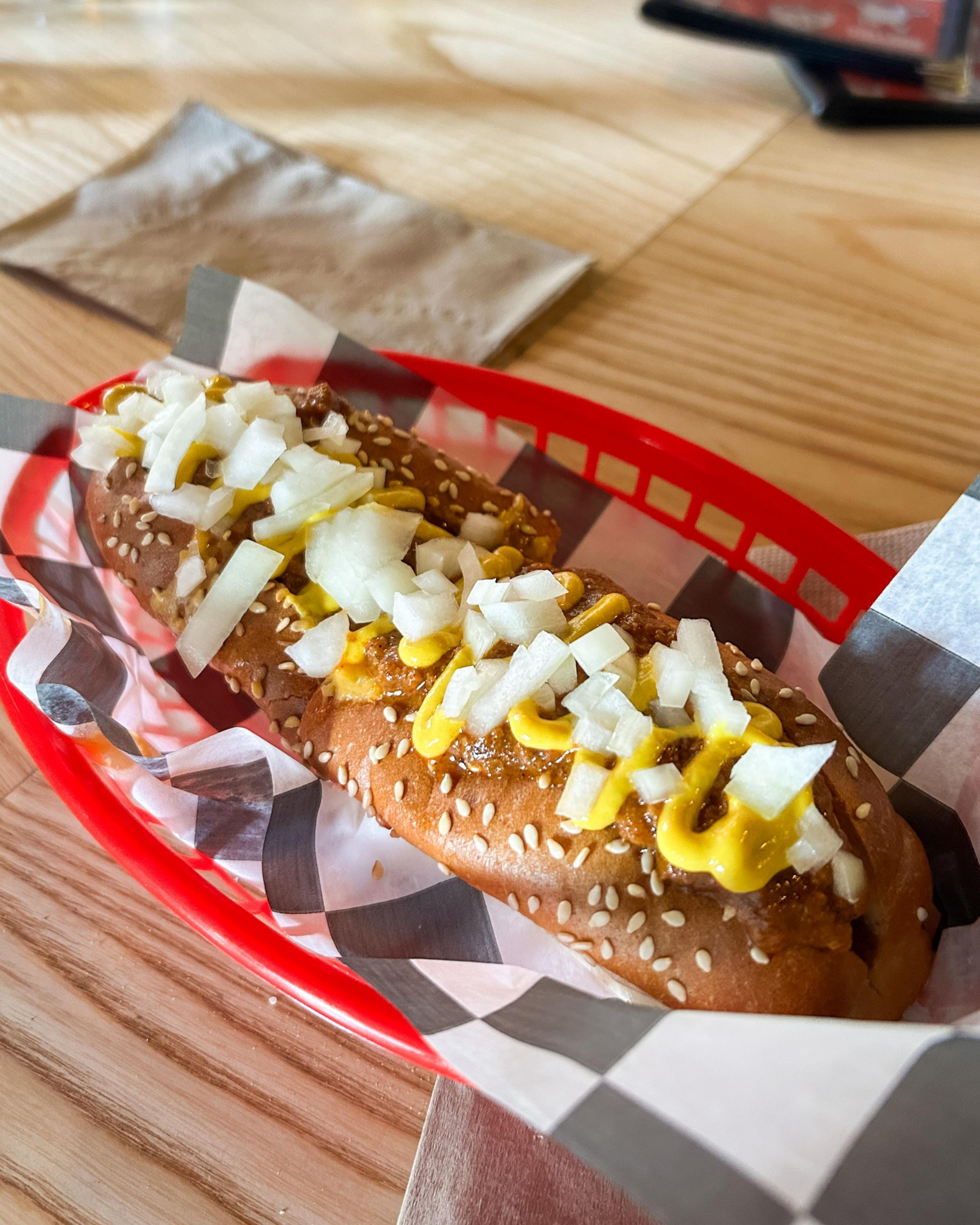 A sesame seed hot dog bun filled with a hot dog, topped with yellow mustard and chopped onions, served in a red basket on checkered paper.