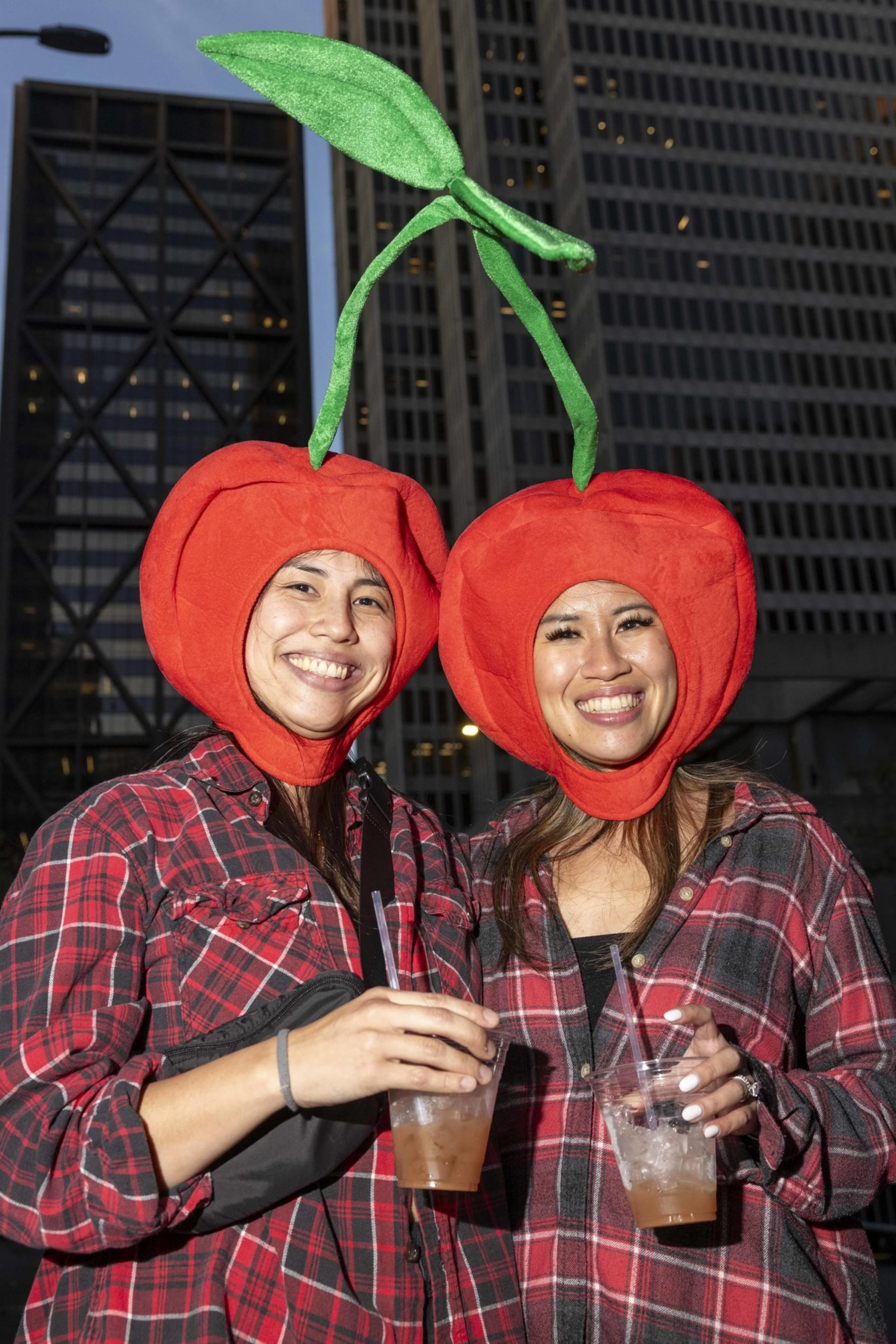 Two people wearing red cherry costumes with green leaves on top, dressed in red plaid shirts, are smiling and holding drinks in city surroundings.