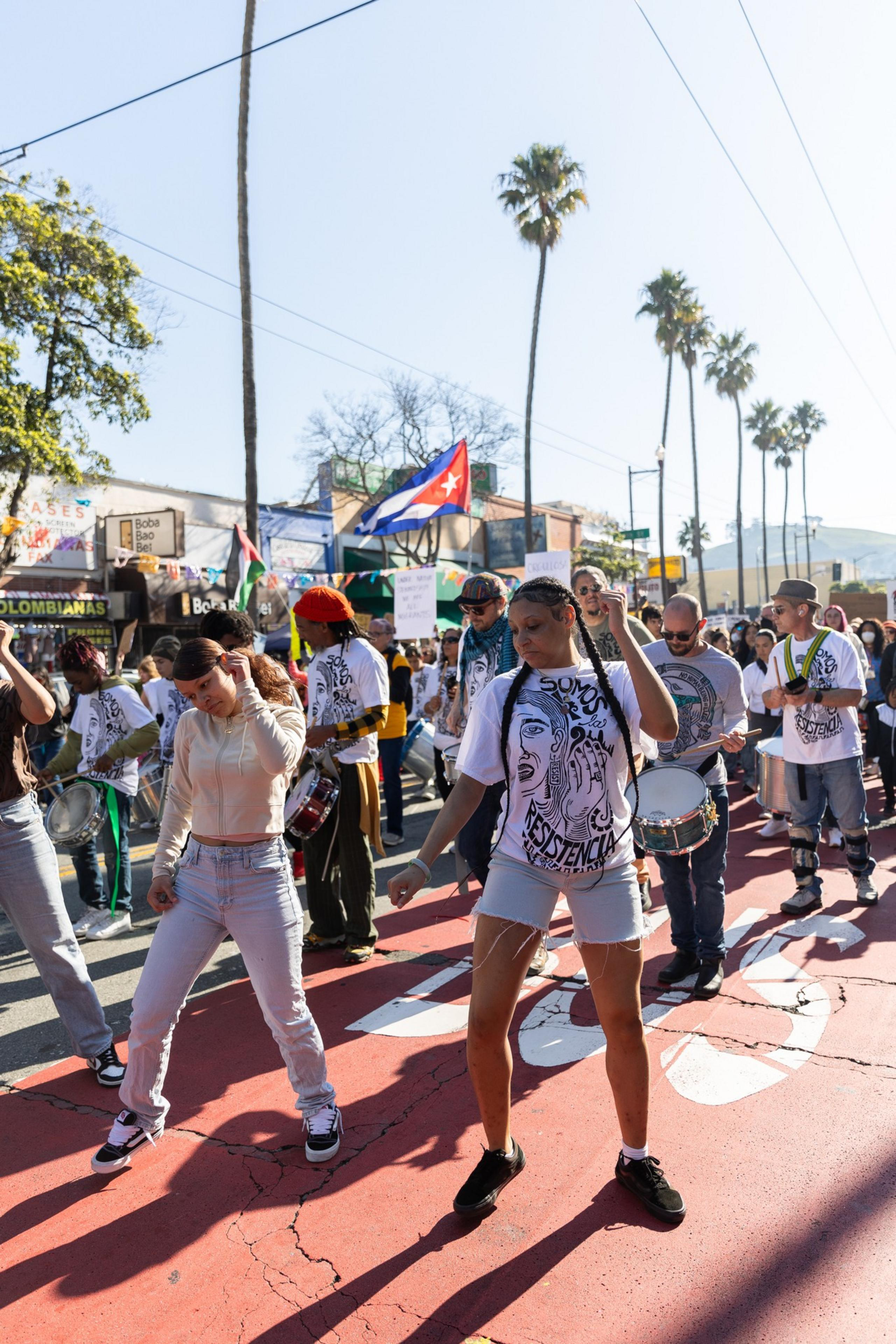 A lively street scene shows people dancing and playing drums. They wear shirts with a graphic design, and a Cuban flag is visible. Tall palm trees line the street.