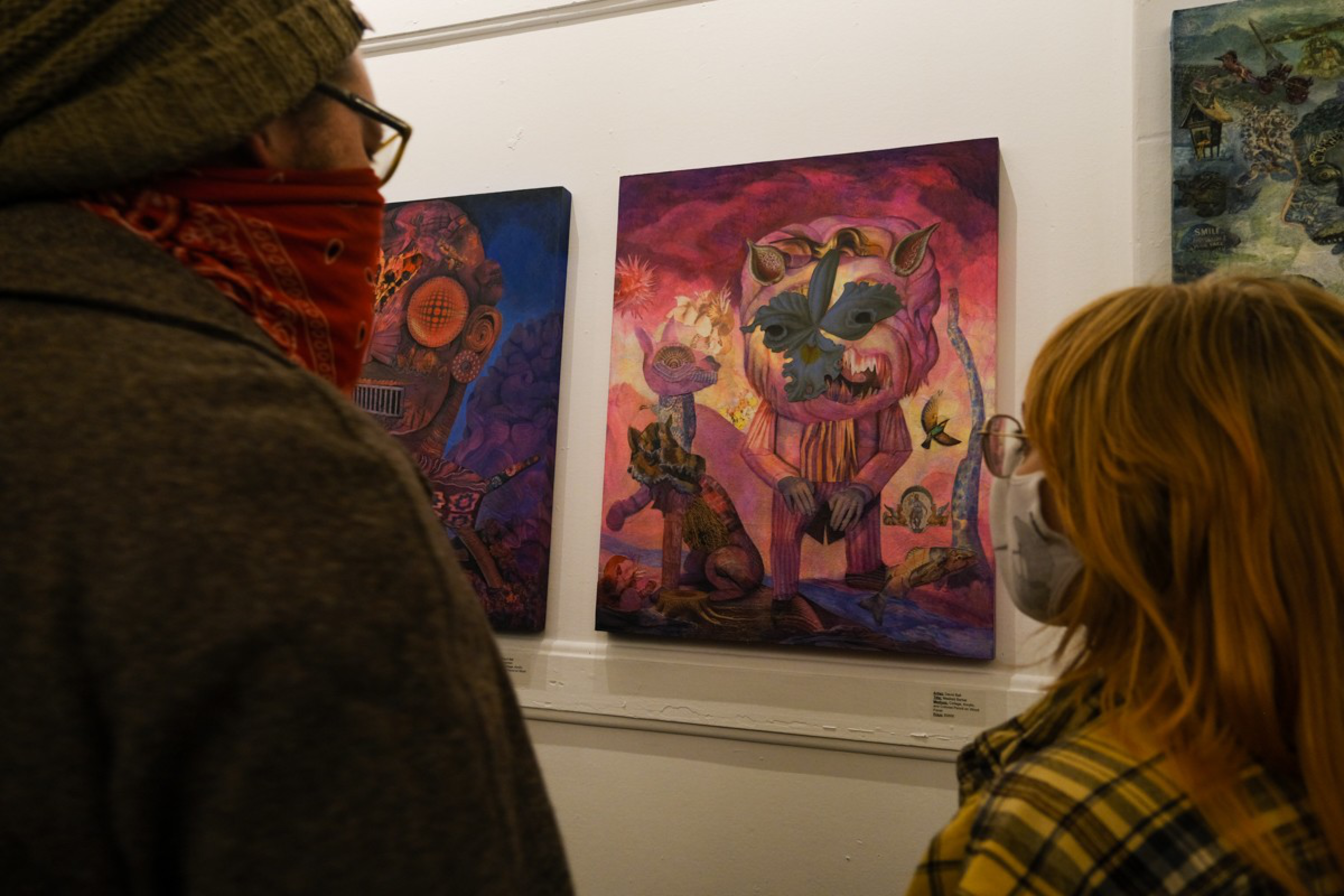Two people wearing masks and glasses closely observe colorful, surreal paintings featuring abstract, fantastical creatures on a white gallery wall.