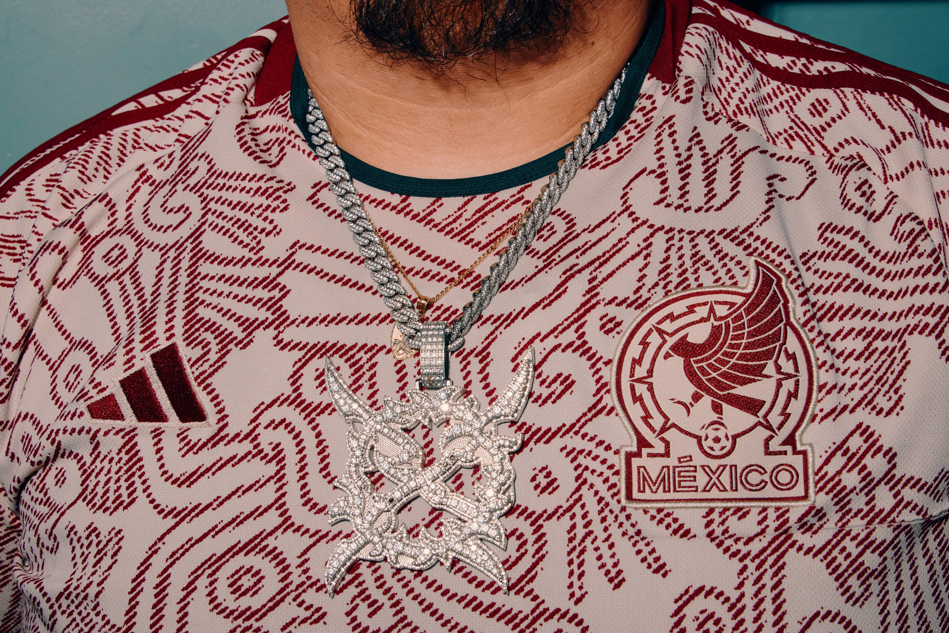 A person wearing a Mexico soccer jersey with red patterns and logos, including an eagle emblem, and two large silver chains with diamond-studded pendants.