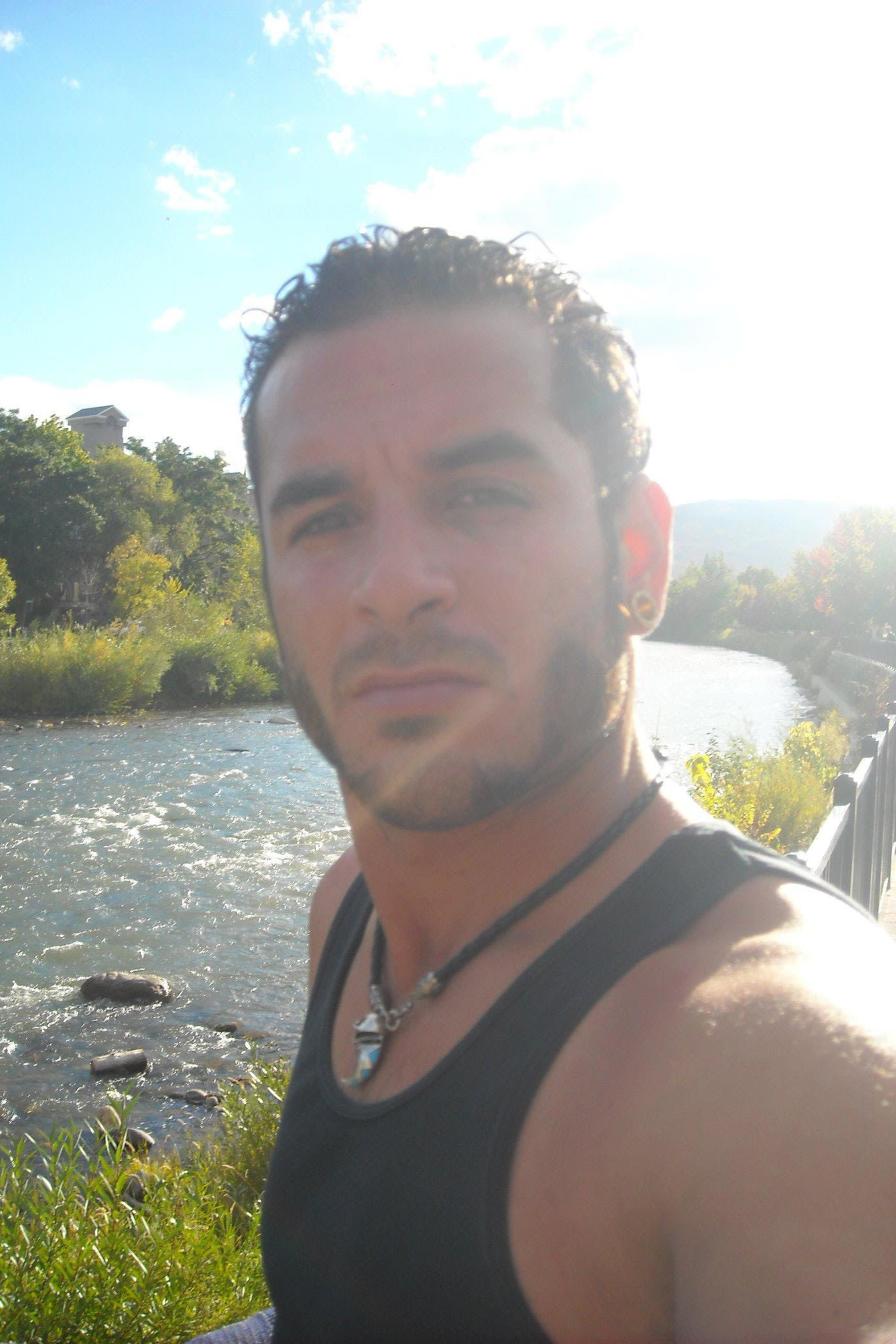 A man with short curly hair and facial hair is taking a selfie by a river on a sunny day. He's wearing a black tank top and necklace; lush greenery surrounds him.