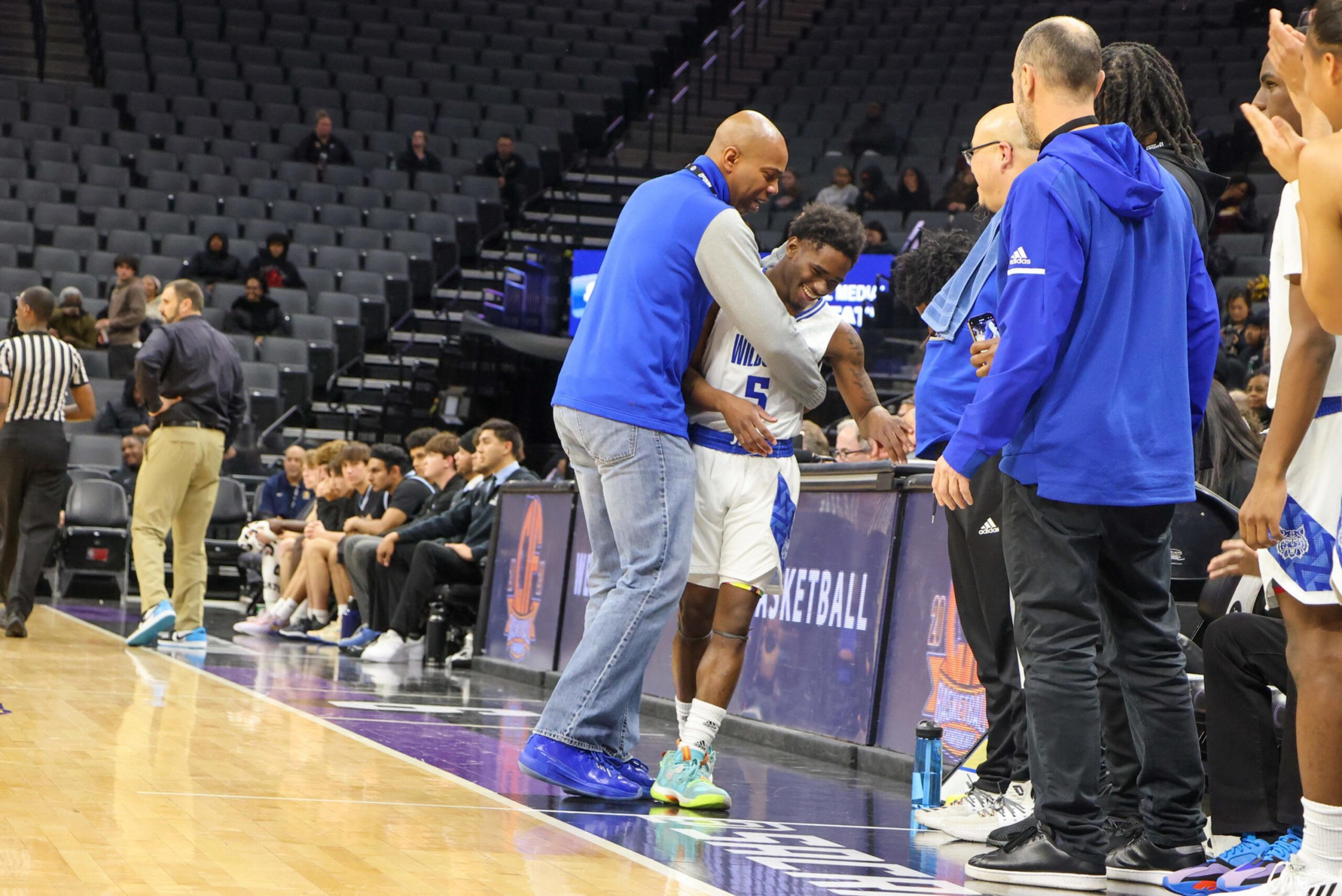 A basketball player is being congratulated and hugged by a coach or staff member near the court, while teammates and coaches watch and applaud.