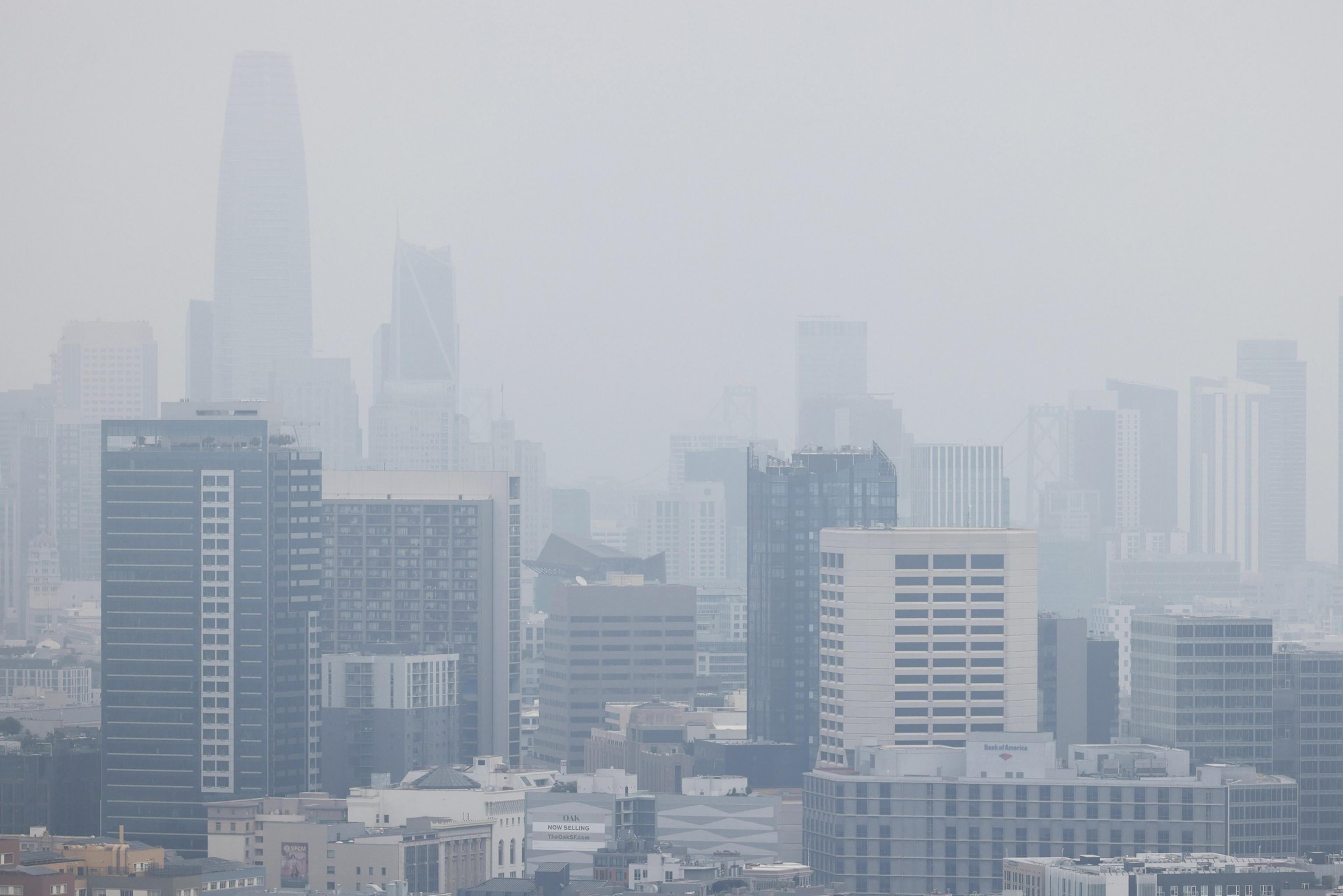 A city skyline is obscured by thick gray smog, making the tall buildings and skyscrapers barely visible through the heavy haze.