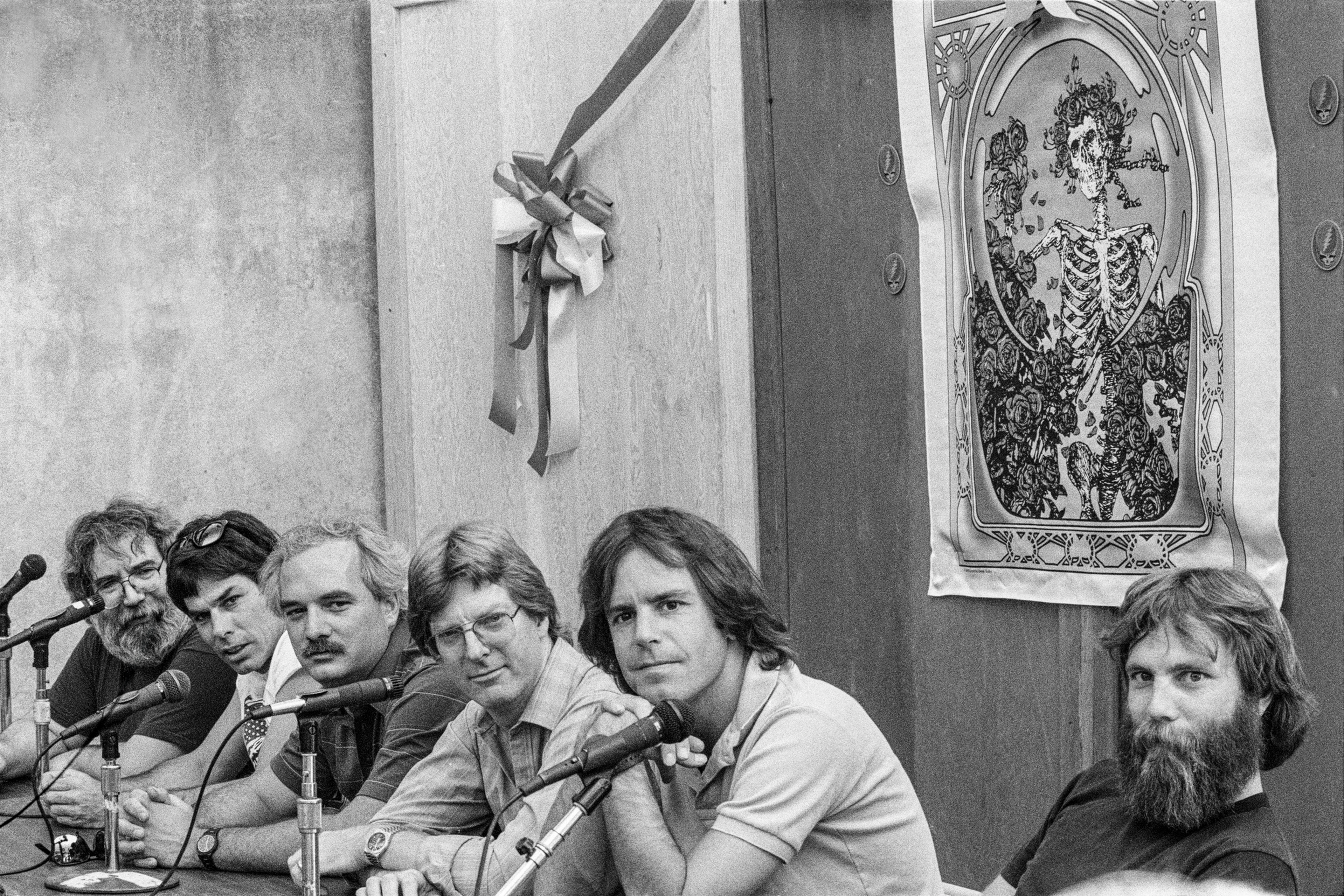 Six men with varying hairstyles and facial hair sit side by side at a table with microphones, against a wall decorated with a ribbon and a skeleton-themed poster.