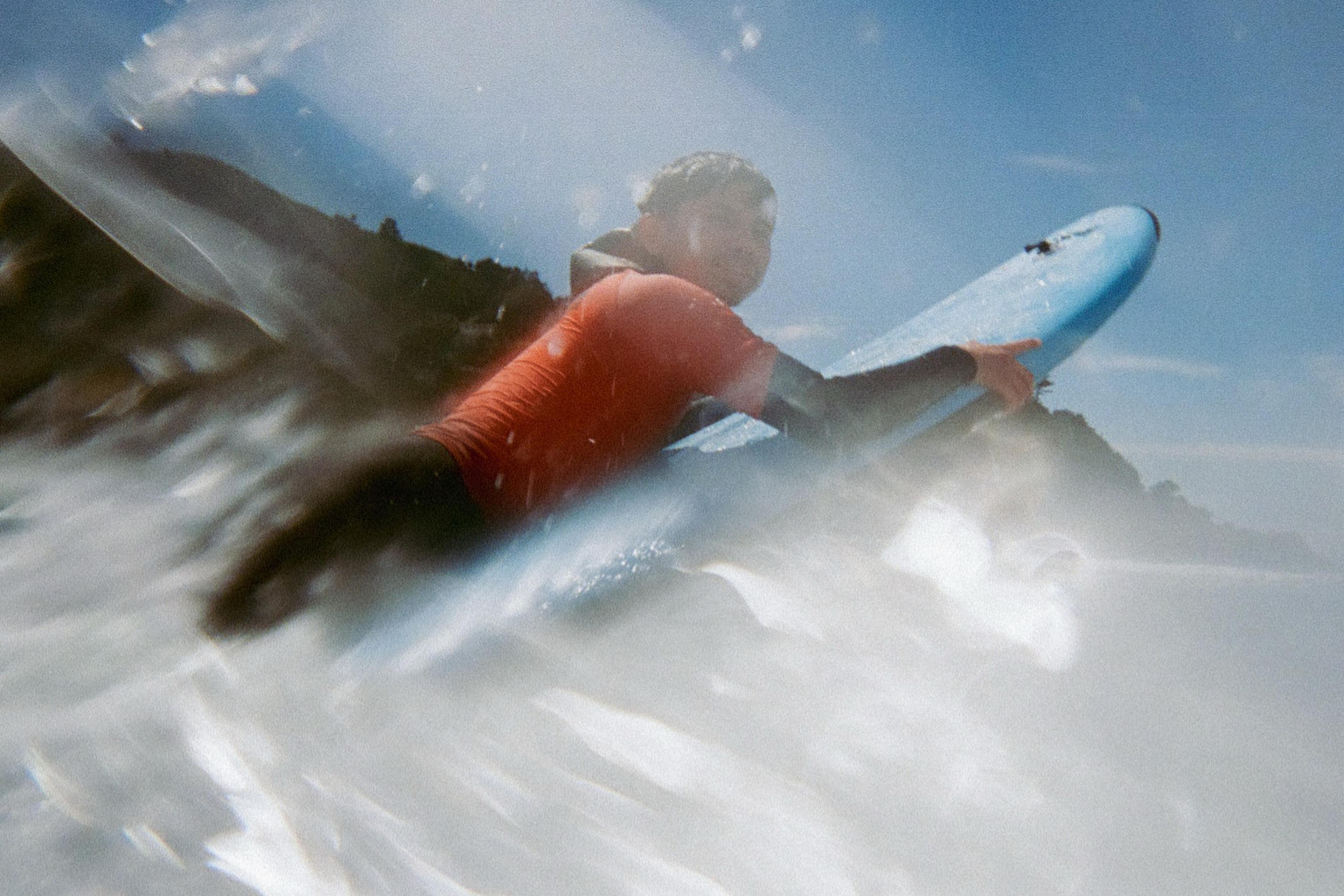 A person wearing an orange shirt is surfing on a blue board amidst frothy waves, with a clear blue sky and distant land in the background.