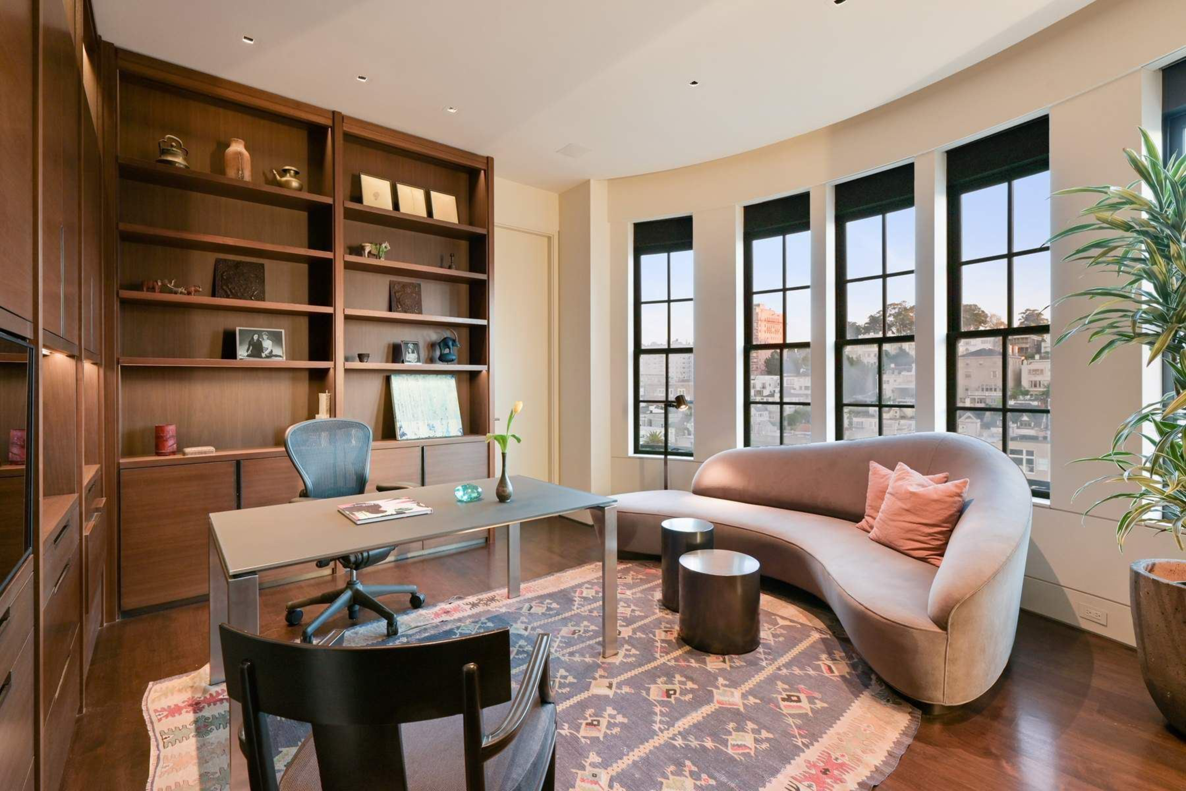 A modern office with a curved beige sofa, three large windows, wooden shelves, a desk with a chair, two round tables, and a patterned rug.
