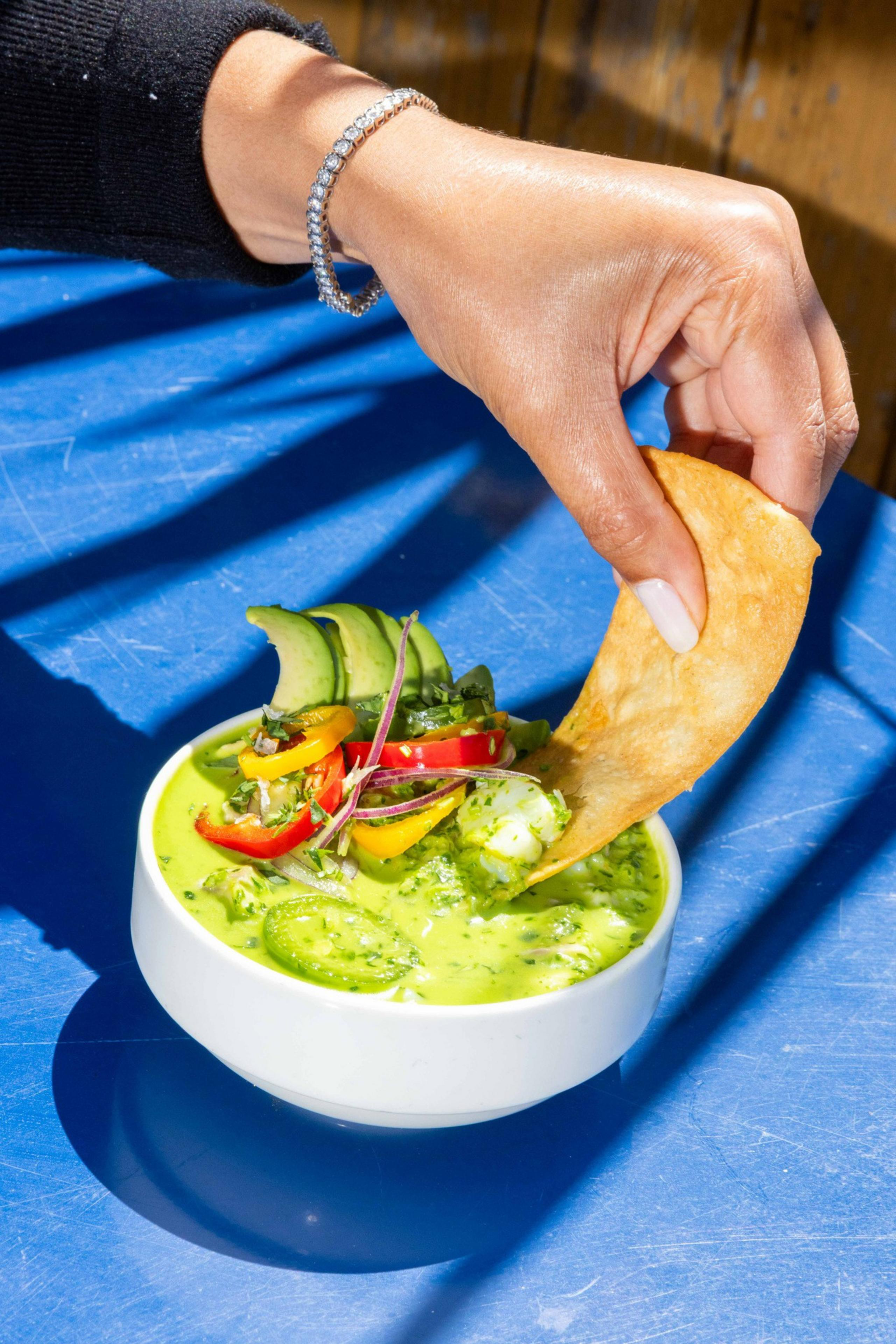 A hand dips a crispy chip into a bowl of creamy green soup, garnished with sliced avocado, jalapeños, and colorful peppers, on a blue table.