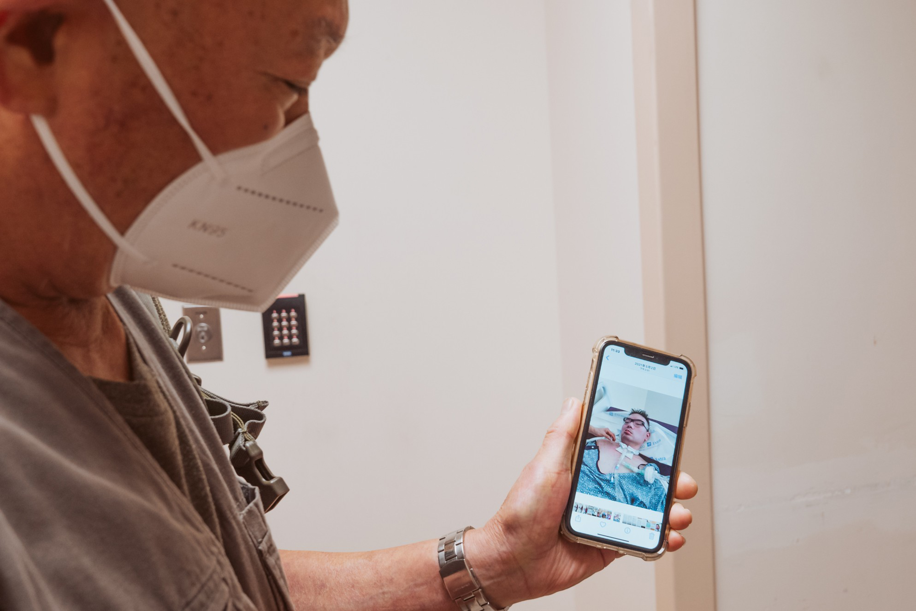 An elderly man wearing a face mask looks at a smartphone displaying a photo of a younger man wearing glasses and a casual shirt.