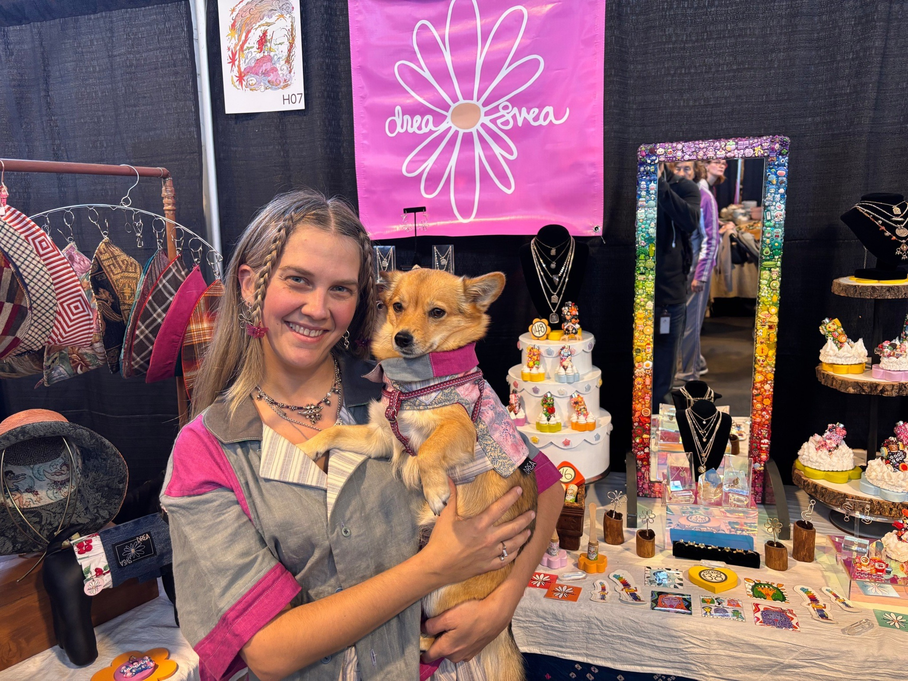 A smiling woman holds a small dog dressed in matching clothes, standing behind a table with colorful jewelry and crafts at a booth with a pink "drea3rea" banner.