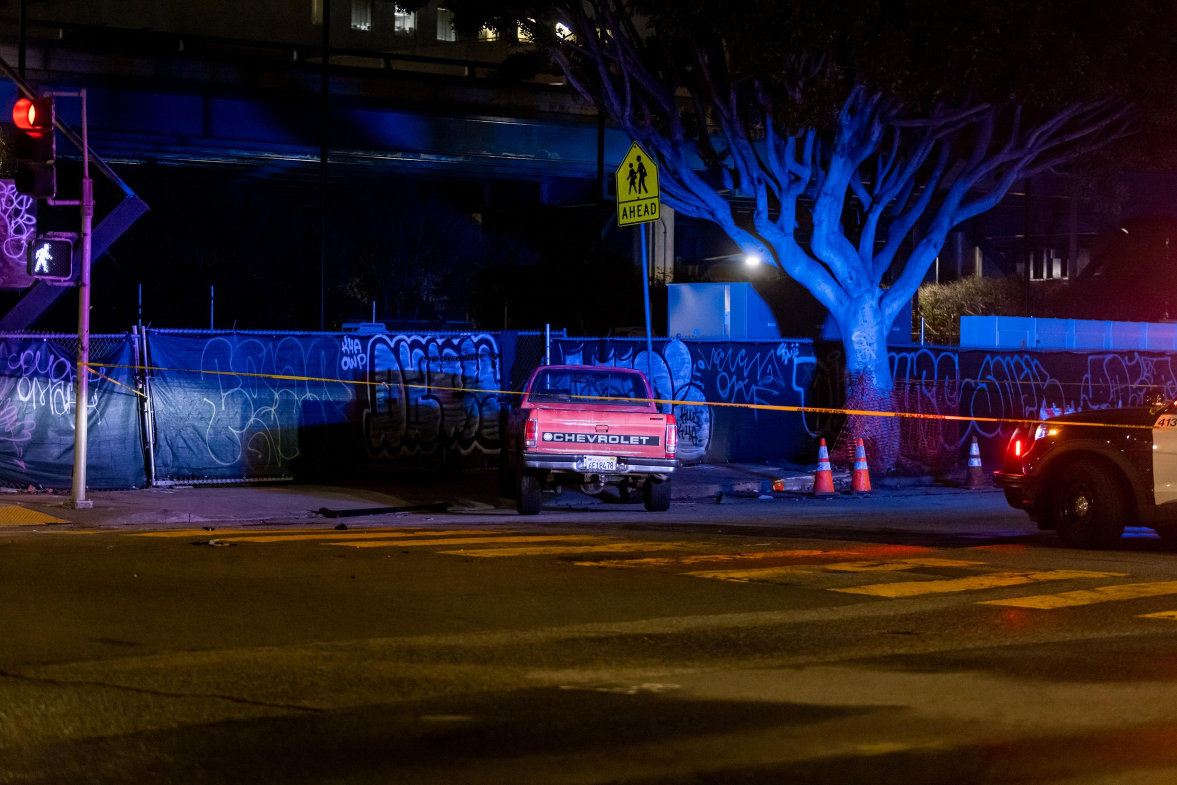 A red Chevrolet truck is parked by a graffiti-covered fence. A police car with flashing lights is nearby, and caution tape surrounds the scene at night.