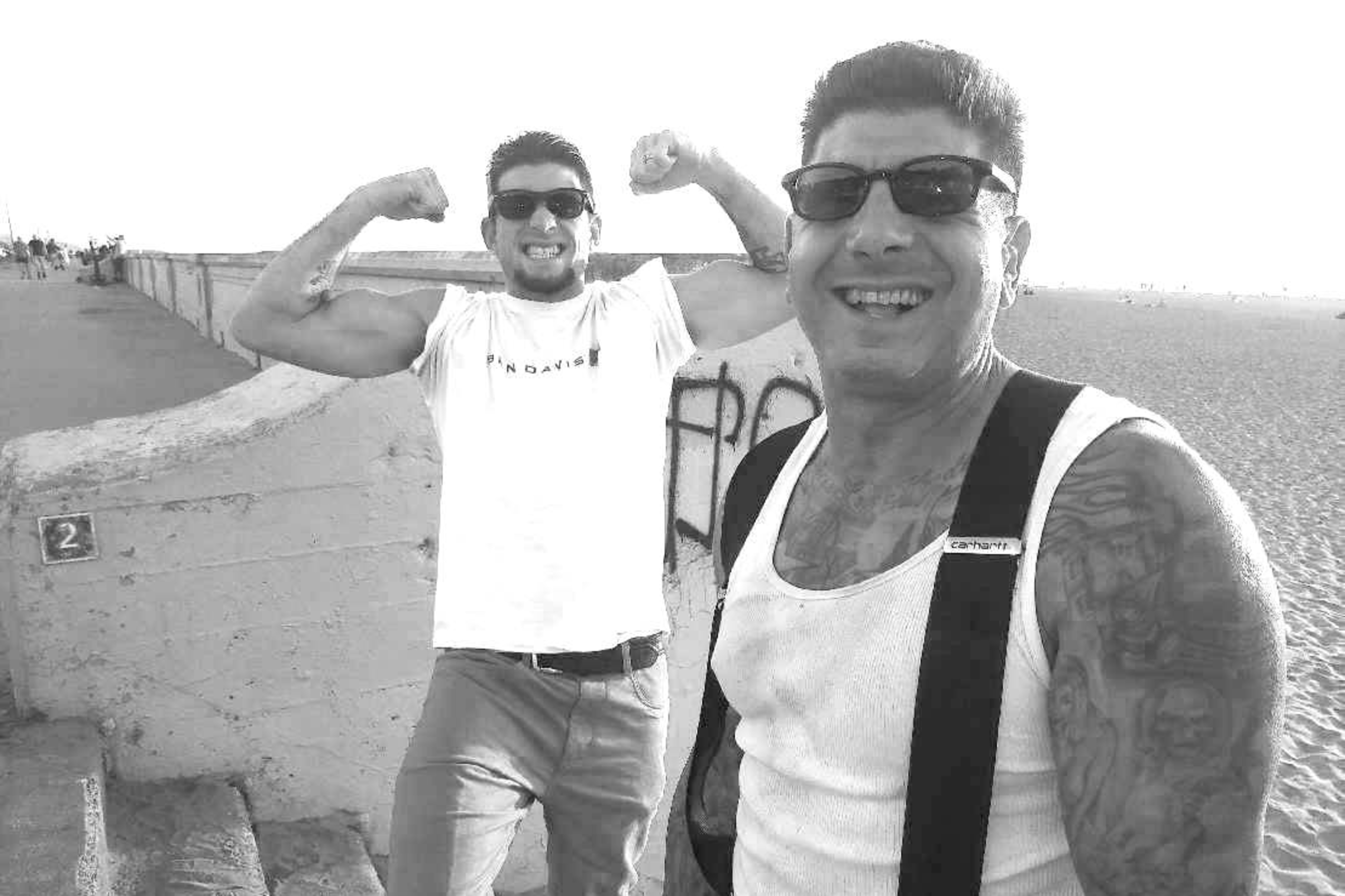 Two men wearing sunglasses smile at the camera; one flexes his muscles while the other shows tattoos on his arms and wears suspenders.