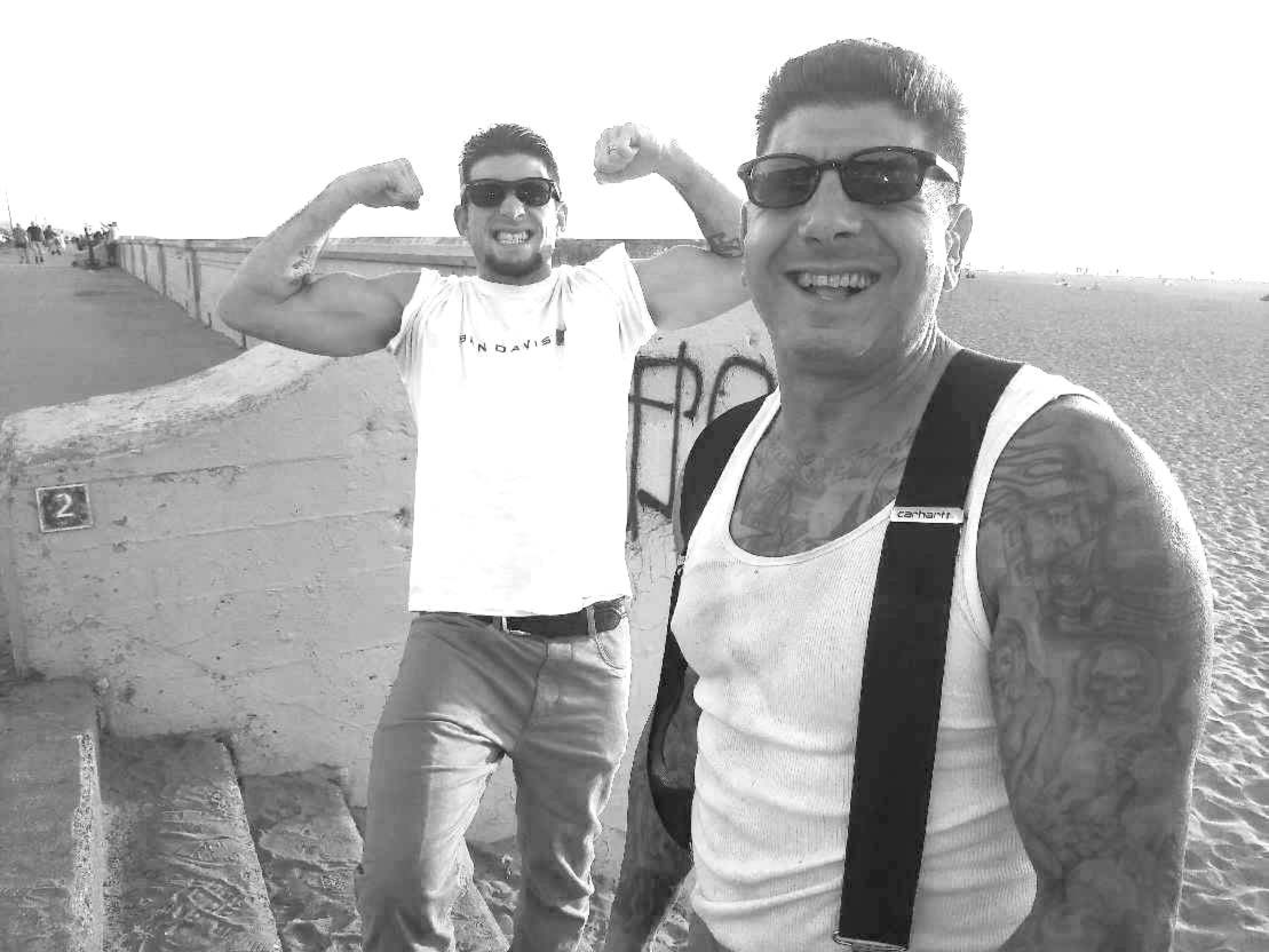 Two men wearing sunglasses smile at the camera; one flexes his muscles while the other shows tattoos on his arms and wears suspenders.
