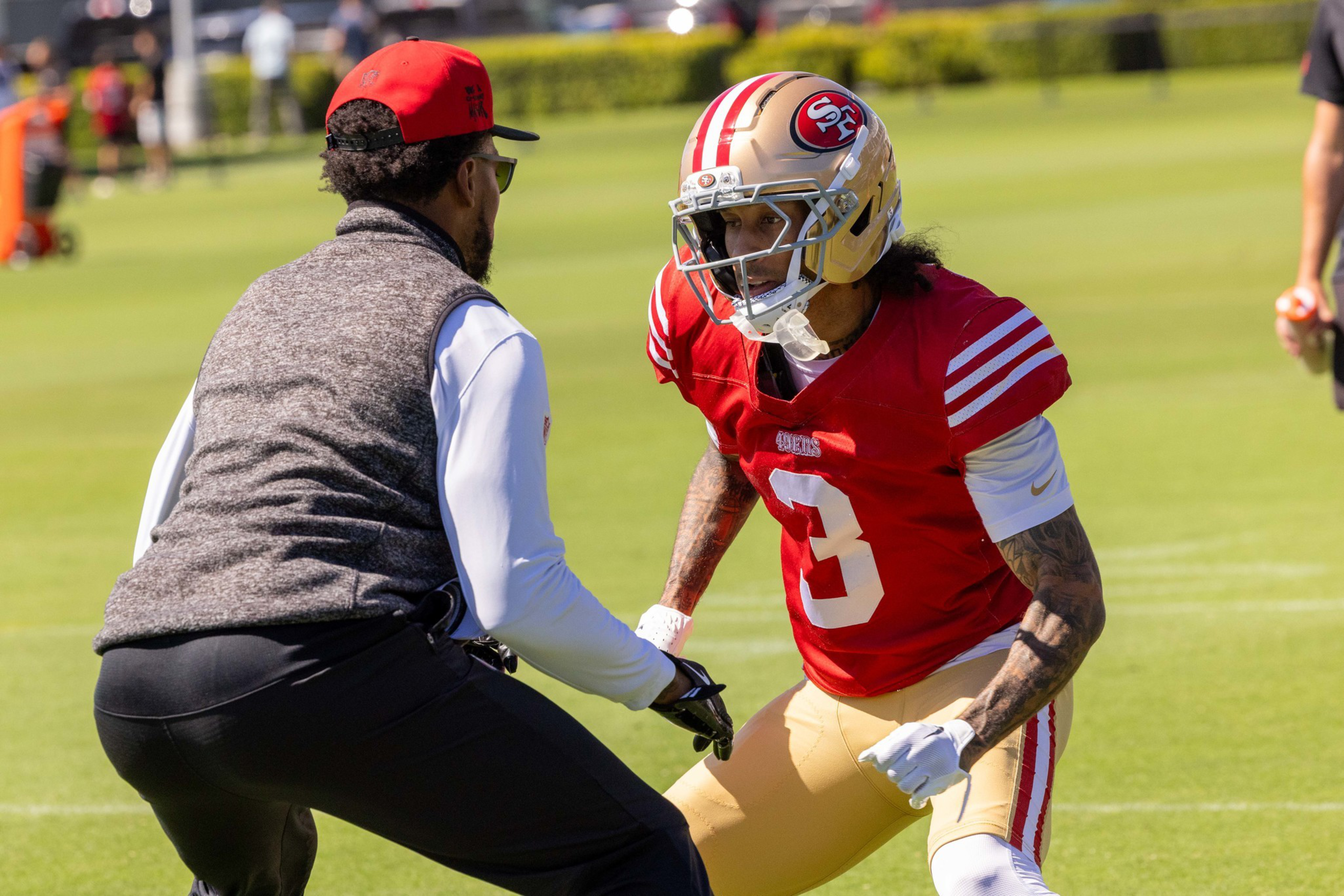 A football player in a red San Francisco 49ers jersey and gold helmet faces a coach wearing a red cap and gray vest on a practice field.