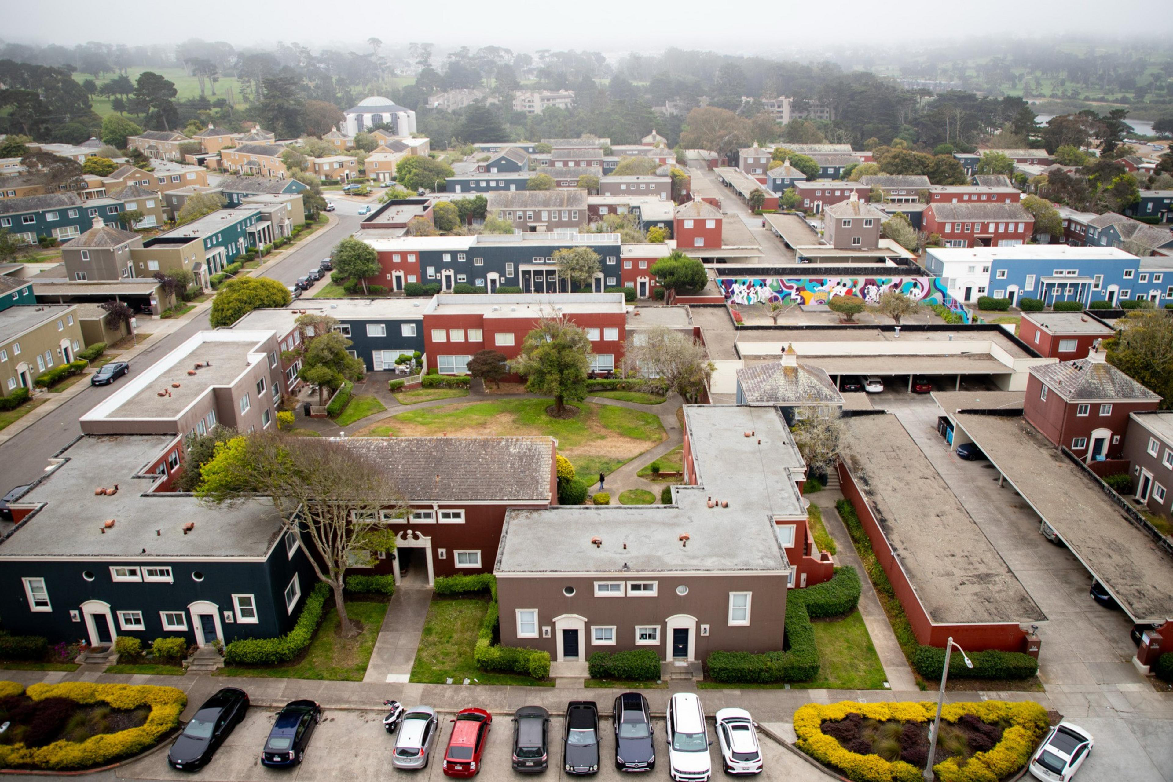 This image shows a suburban neighborhood with multiple modern houses in muted colors, surrounded by roads and greenery. There are parked cars and a colorful mural.