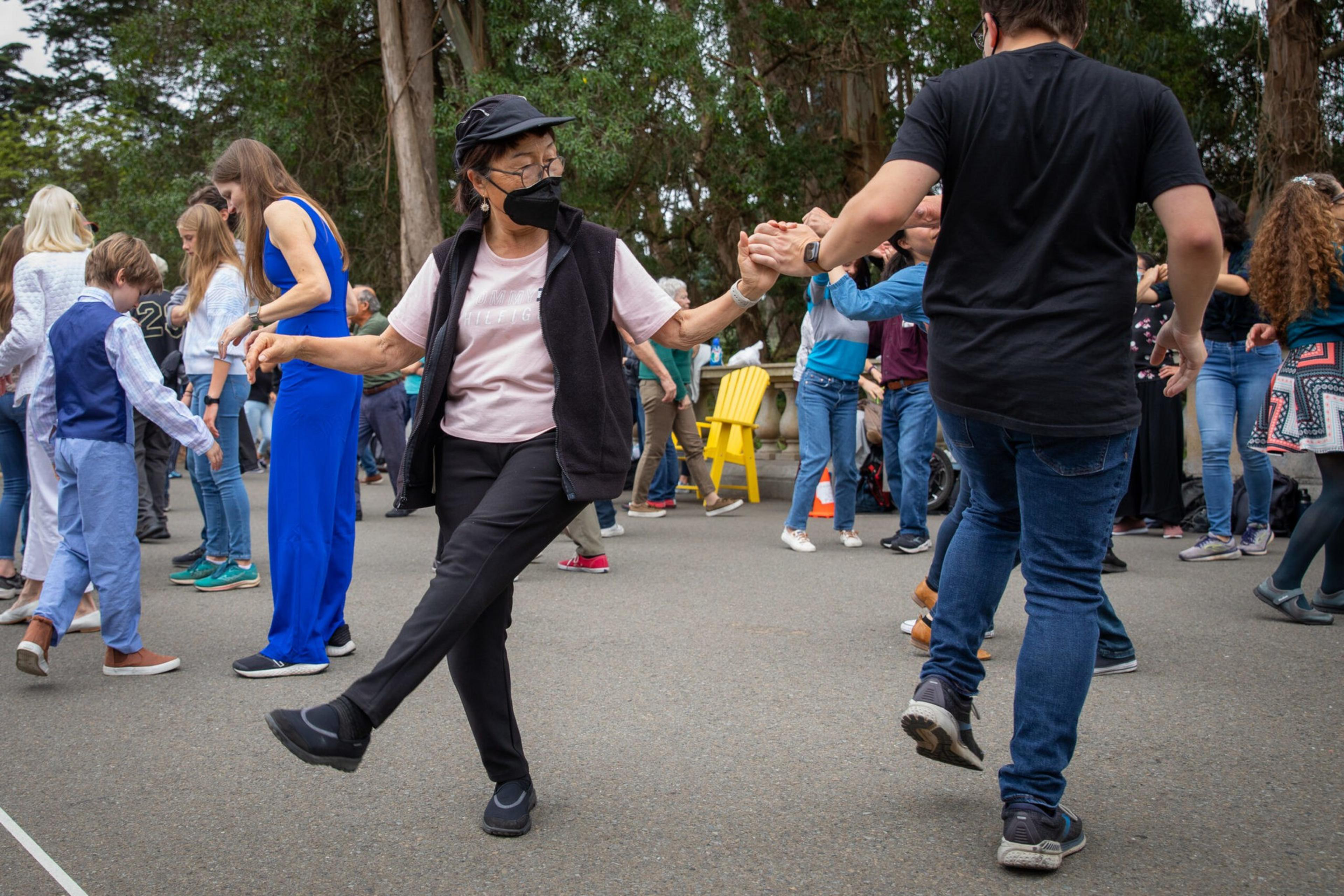 People of various ages are dancing outdoors, holding hands, with trees and a yellow chair visible in the background.