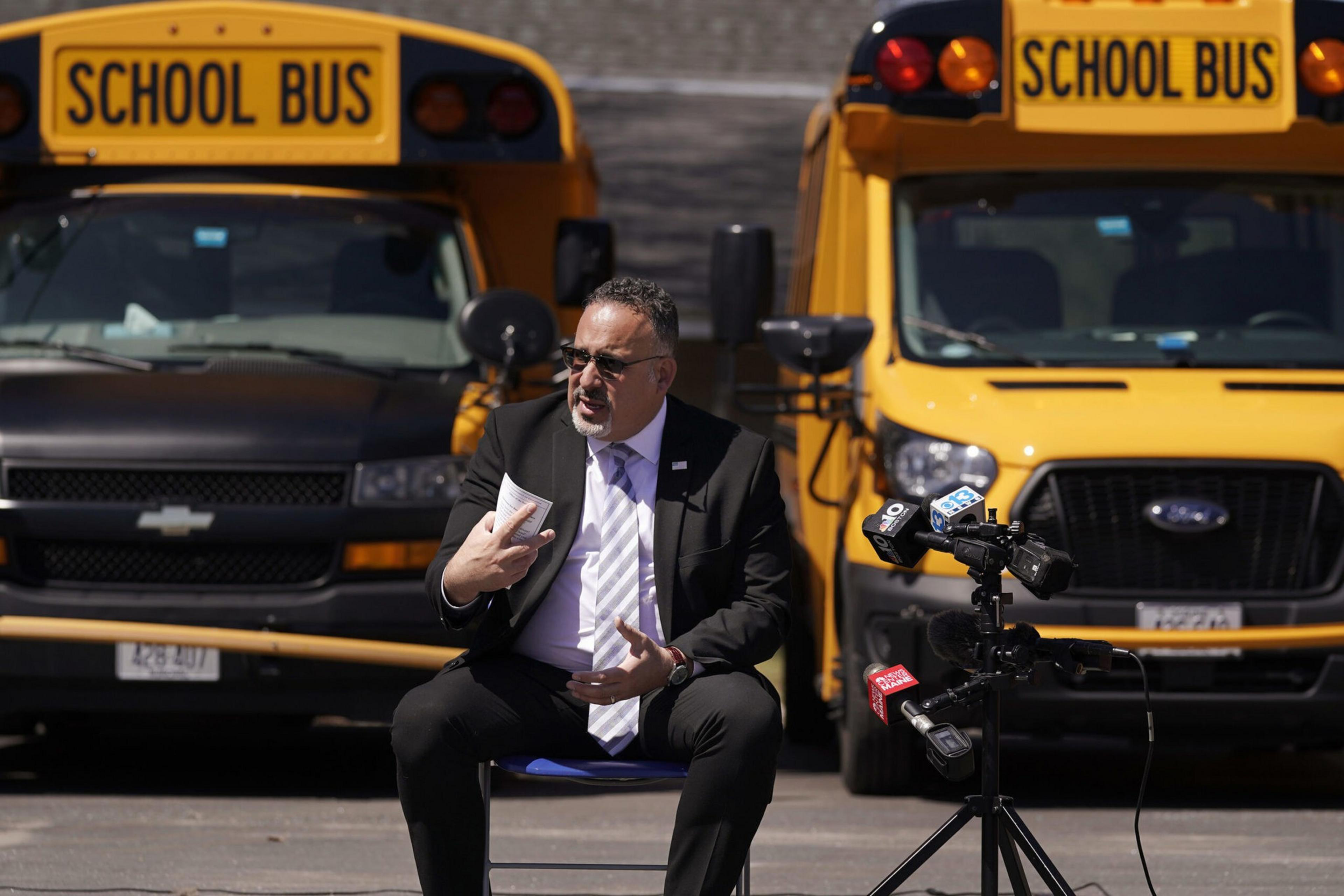 A man in a suit and tie sits speaking in front of two yellow school buses, with microphones set up nearby.