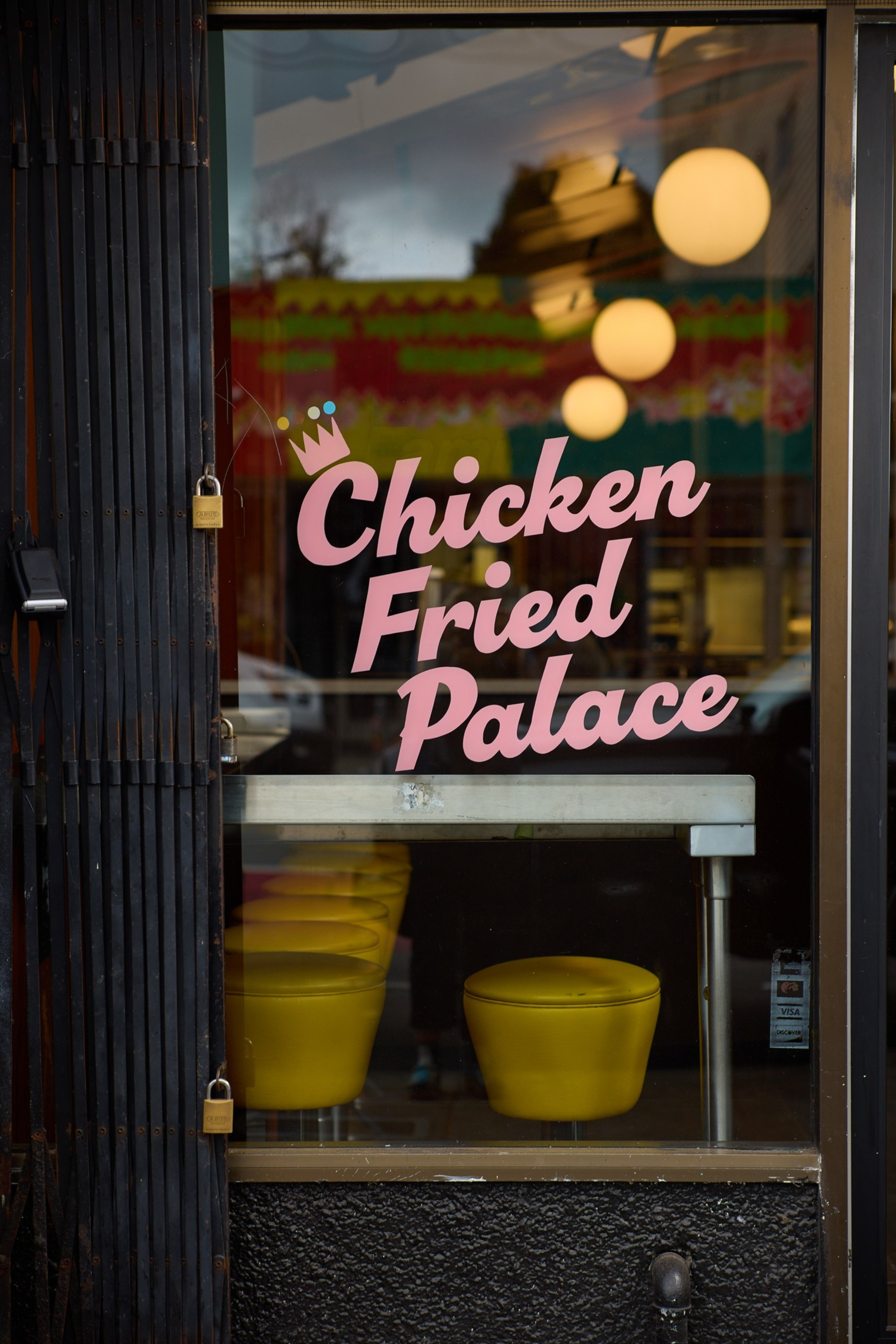 A glass door with pink lettering reading “Chicken Fried Palace” shows a row of yellow stools inside and round hanging lights reflecting on the glass.