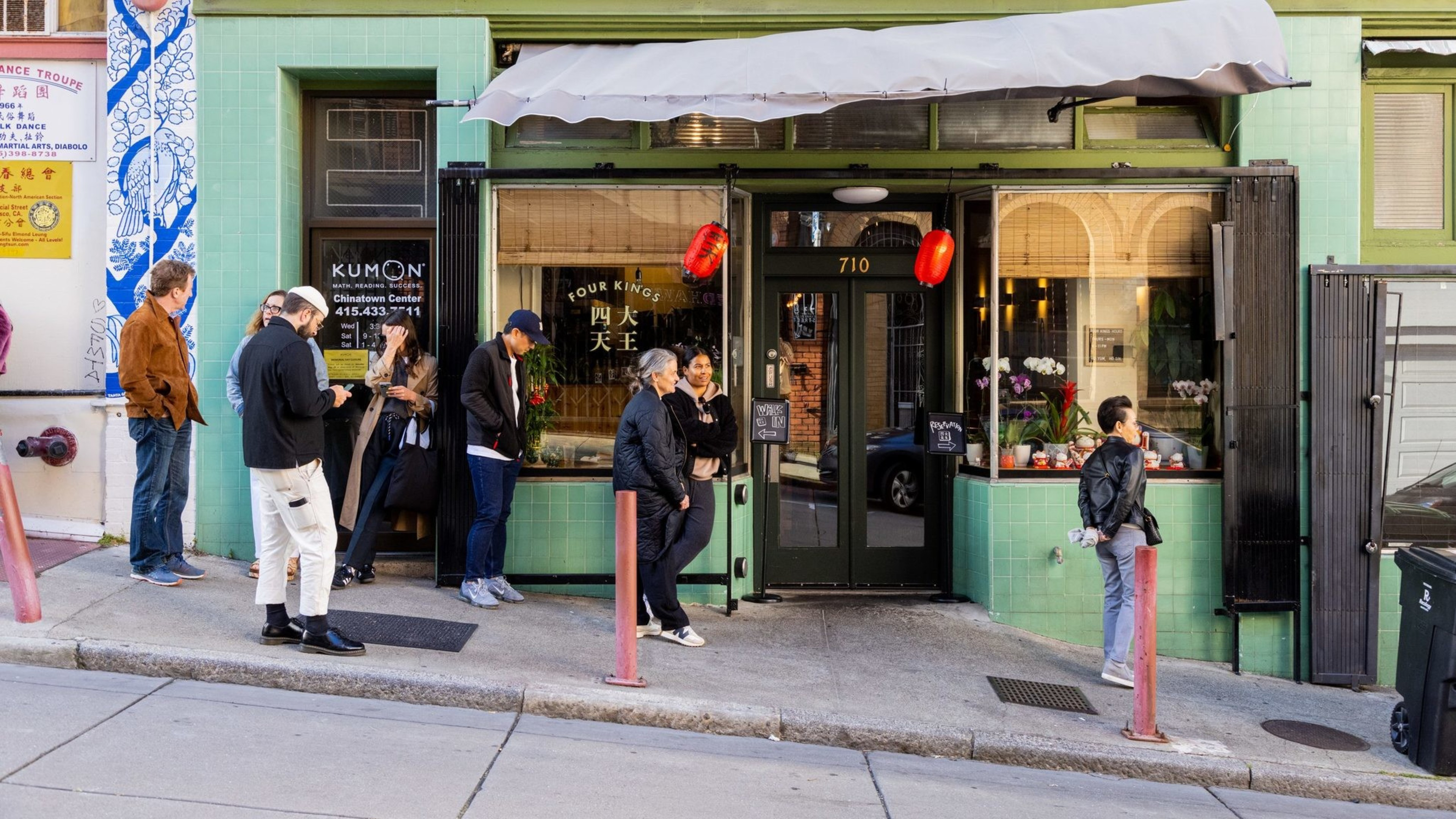 People are lined up outside a small, mint-green restaurant with red lanterns and plants in the window. Some chat while others look at their phones.
