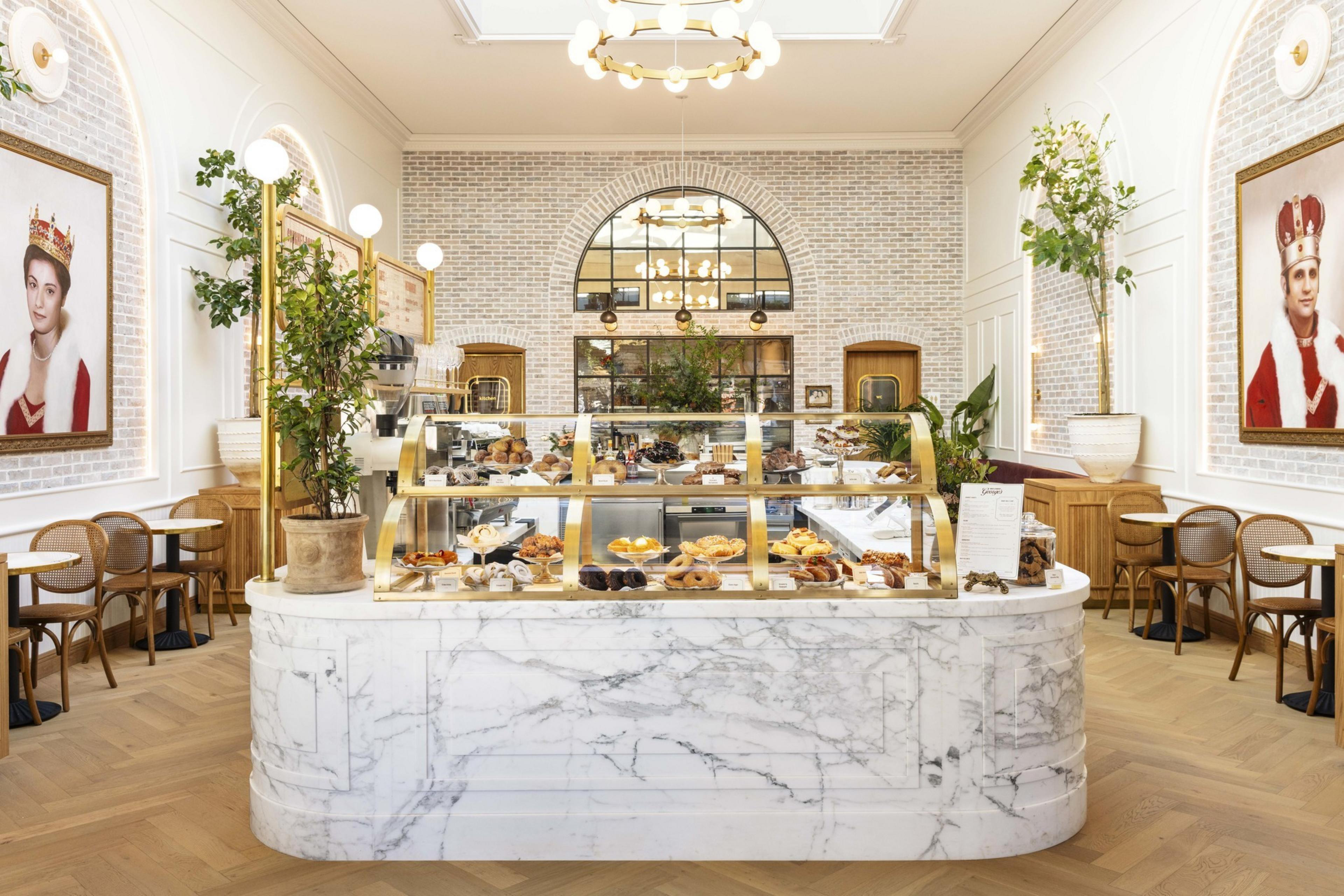 The image shows a bright bakery with a marble counter and glass display filled with pastries. Elegant seating, indoor plants, and large portraits adorn the space.