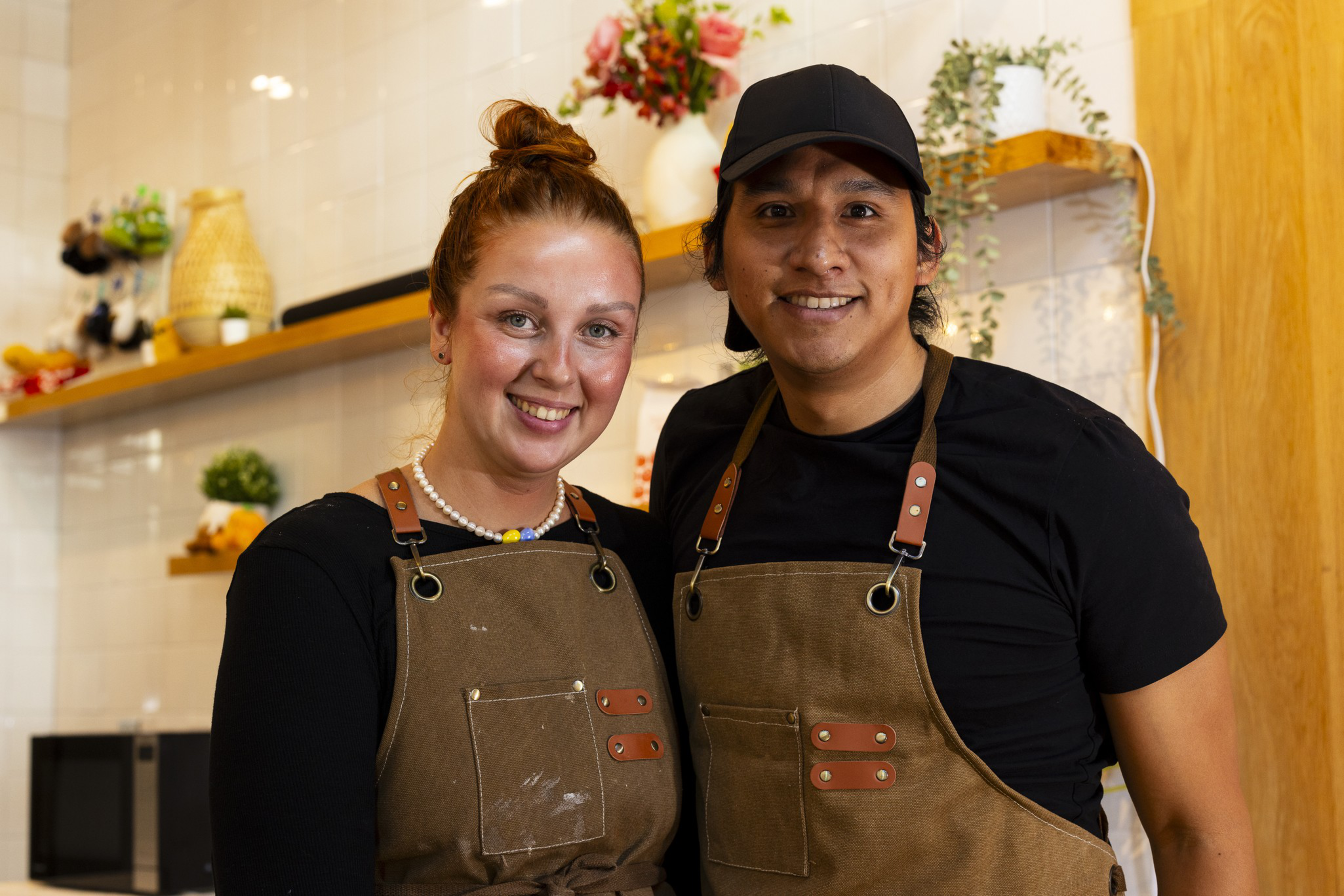 A smiling man and woman wearing matching brown aprons stand close together in a warmly lit kitchen with shelves holding plants and decor.