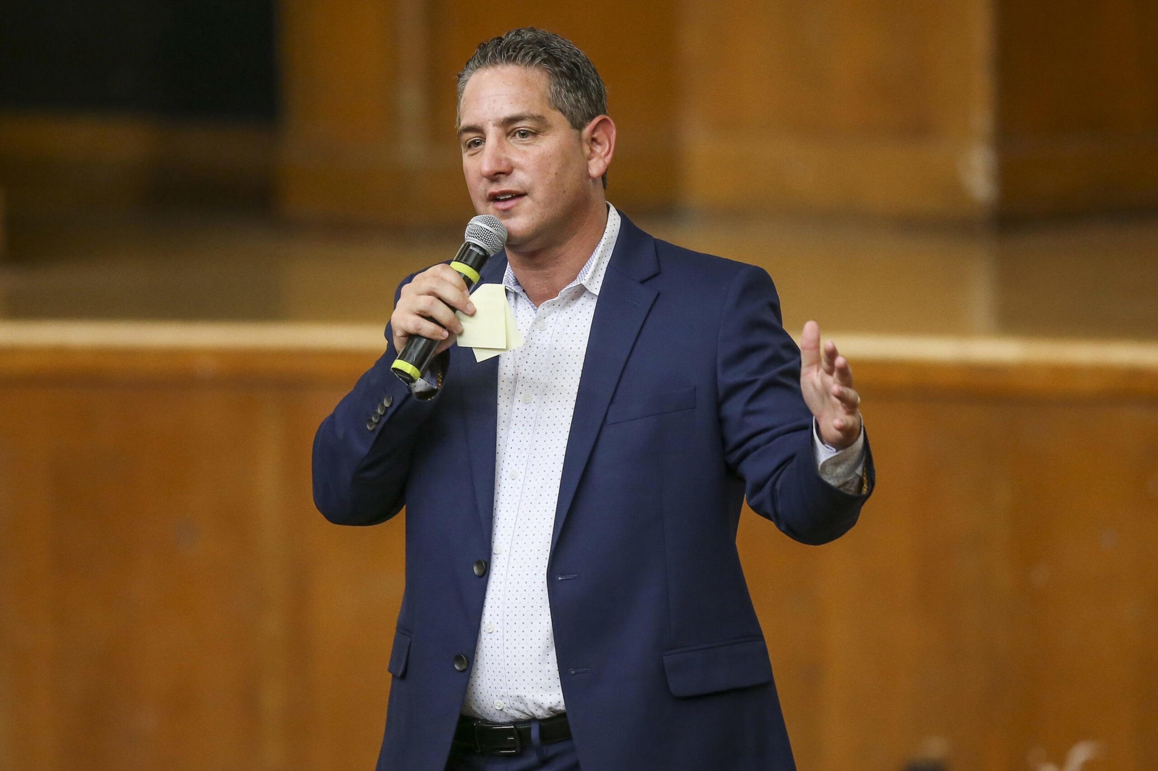 A man in a dark blue suit speaks into a microphone, gesturing with his other hand, standing in front of a blurry wooden background.