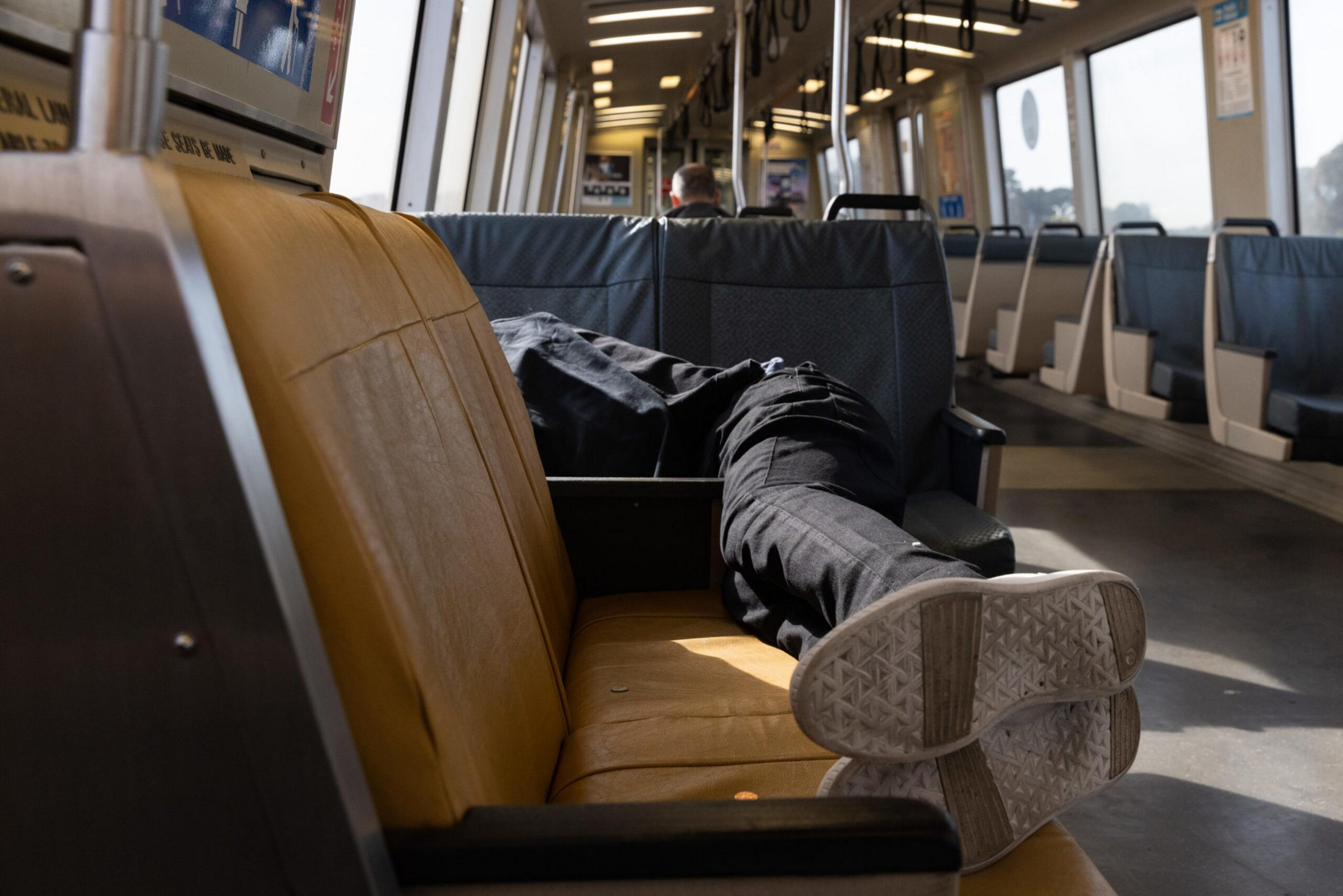 A person lies down across seats on a mostly empty train, wearing black pants and white sneakers, with their feet facing the camera.