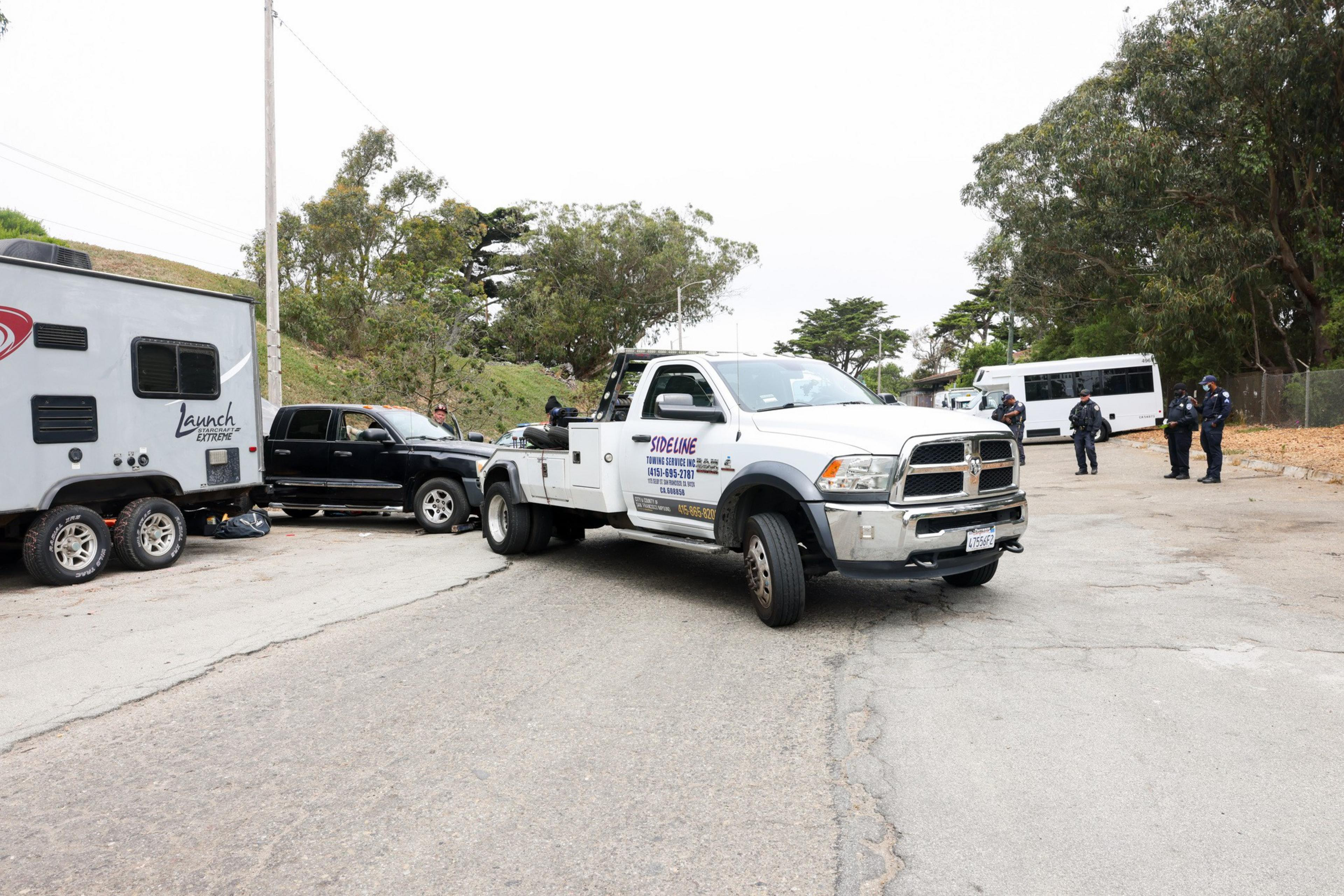 The image shows a white tow truck, a black pickup, and a travel trailer on a street lined with trees. Several police officers stand nearby, along with a white van.