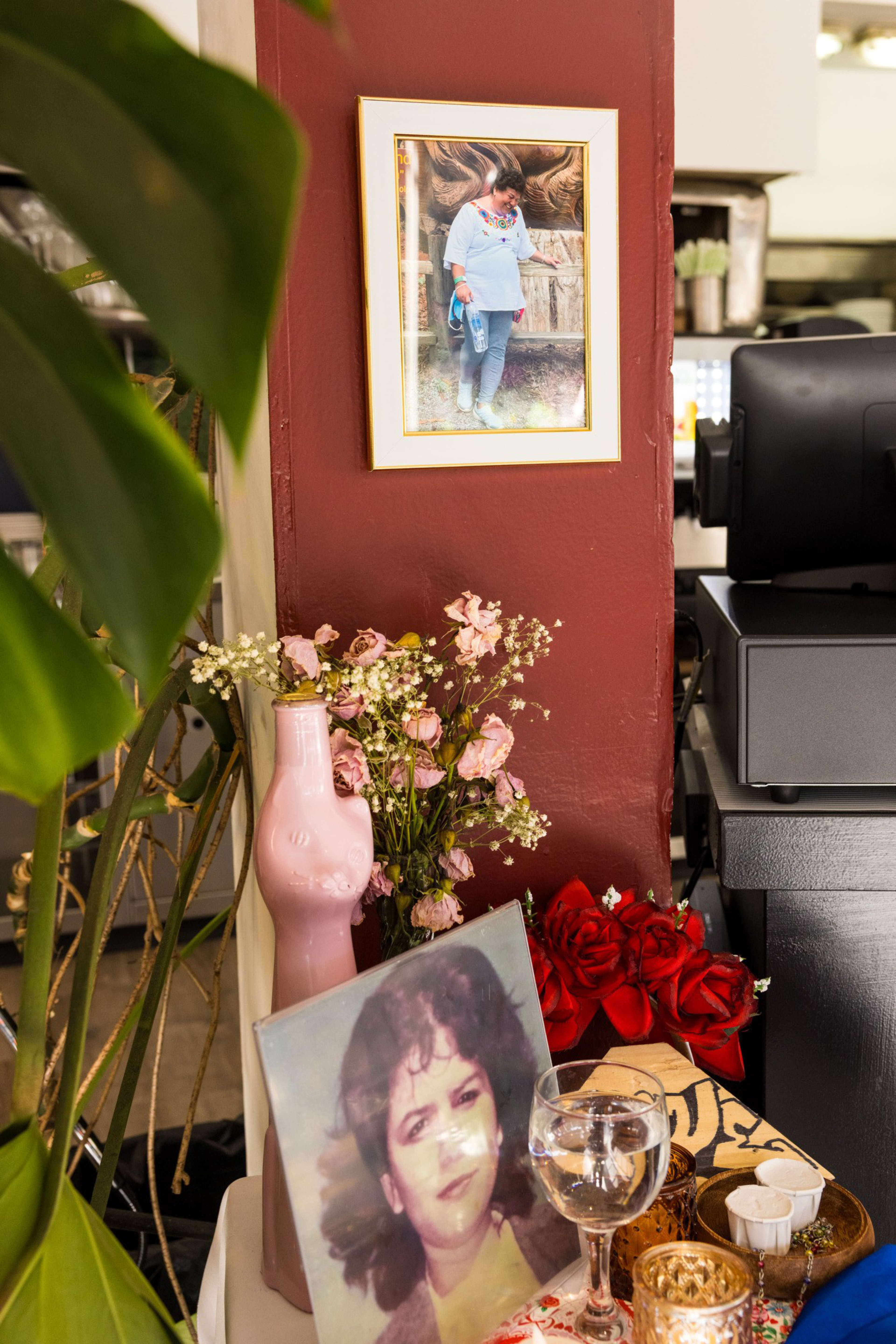 A framed photo of a woman is on a reddish wall. Below, there's a pink vase with dried flowers, a portrait photograph, red roses, and a glass of water.