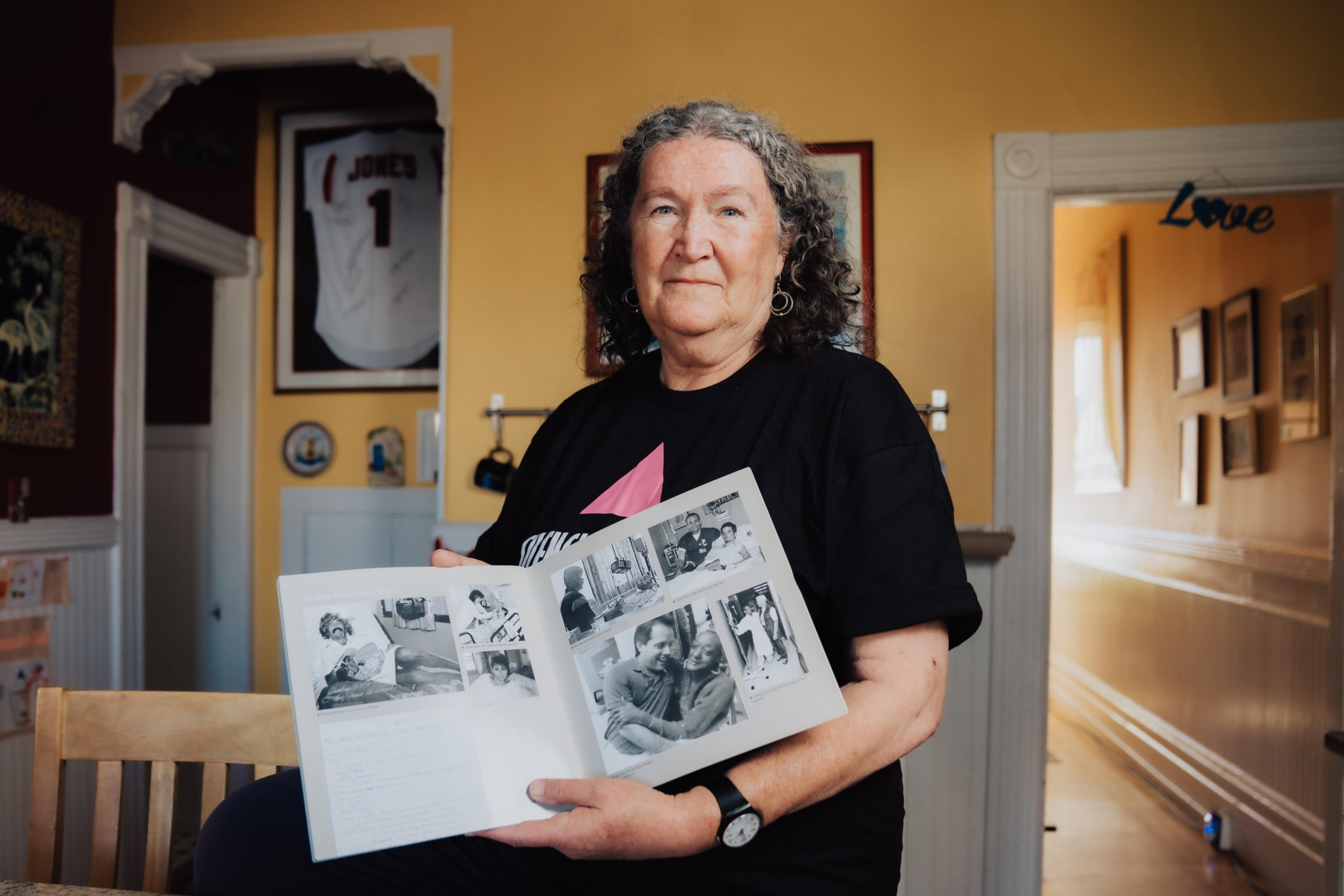 A woman with curly gray hair holds an open scrapbook displaying black-and-white photos. She stands in a warmly lit room with framed items on the walls.