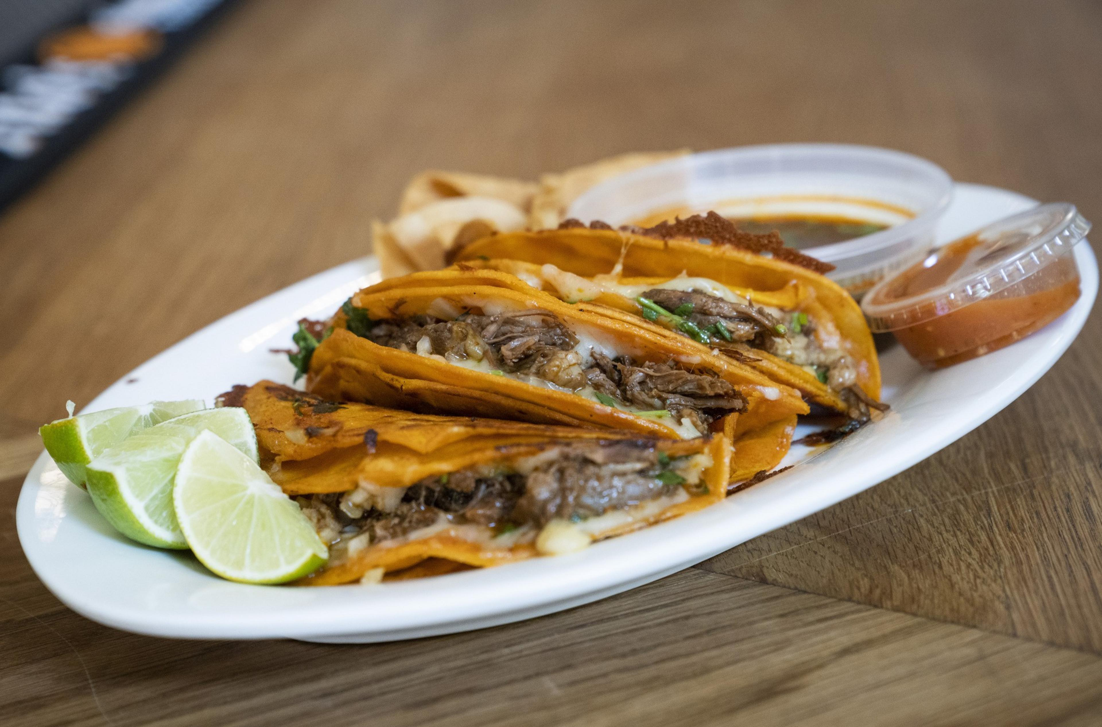 Three birria tacos with melted cheese and beef are on a white plate, accompanied by lime wedges, crispy tortilla chips, and small containers of sauce.