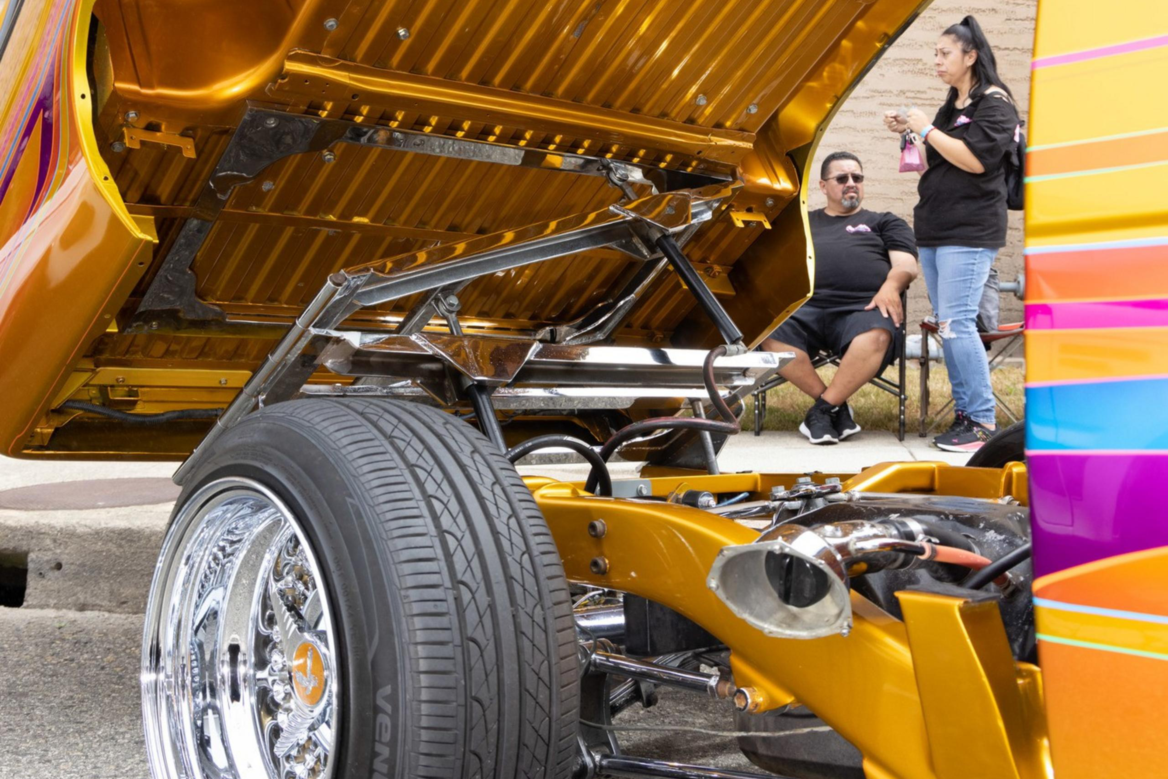 A brightly colored car with its hood open, showcasing intricate detailing. A man and woman conversing nearby.