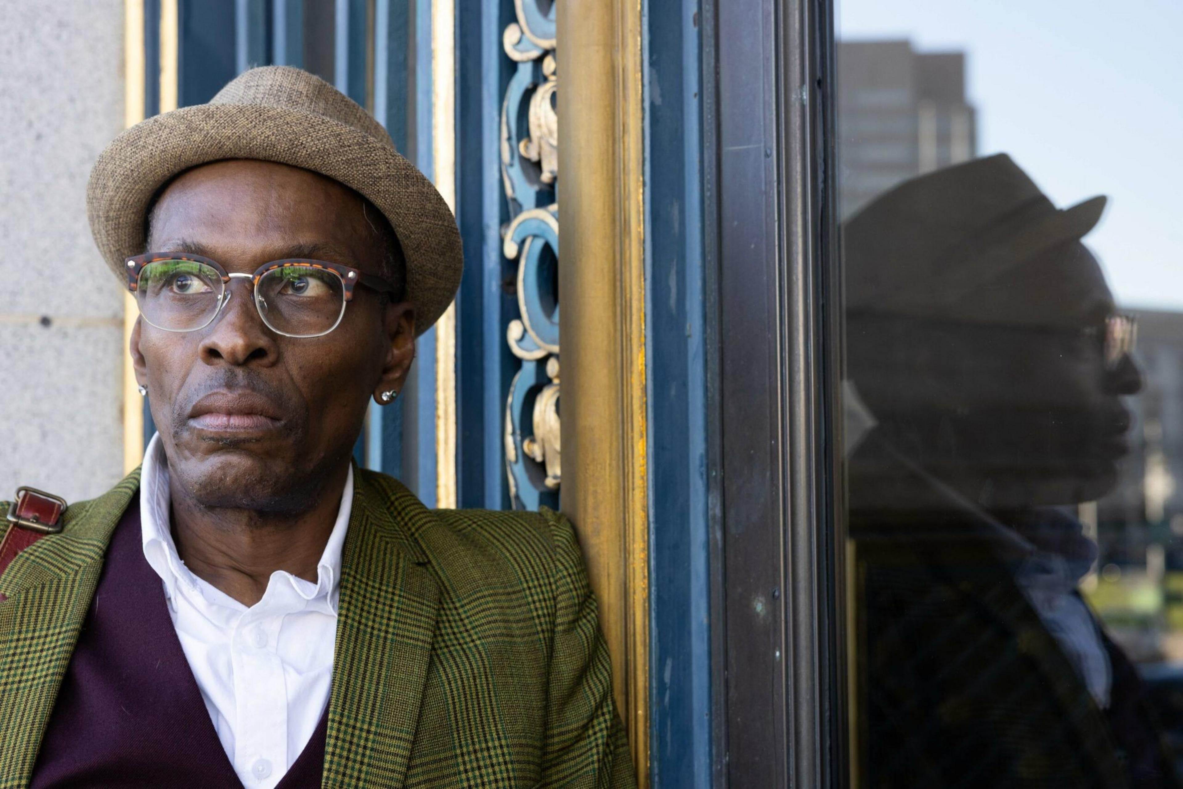 A man wearing glasses, a tan fedora, and a green plaid jacket stands next to a window reflecting his thoughtful profile.