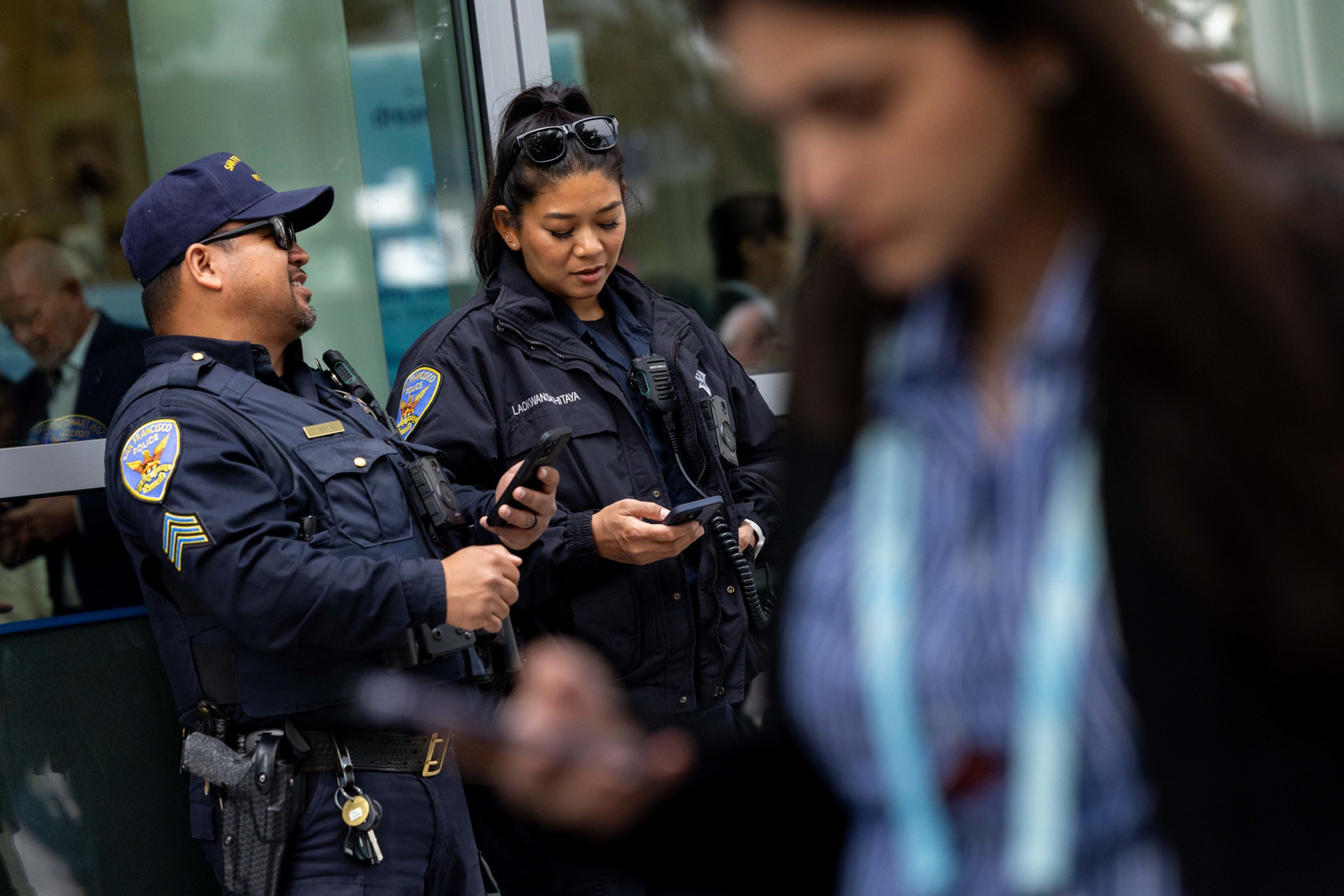 Two police officers in uniform stand outside, one smiling and the other focused on their phone, while a person in the foreground also looks at a phone.