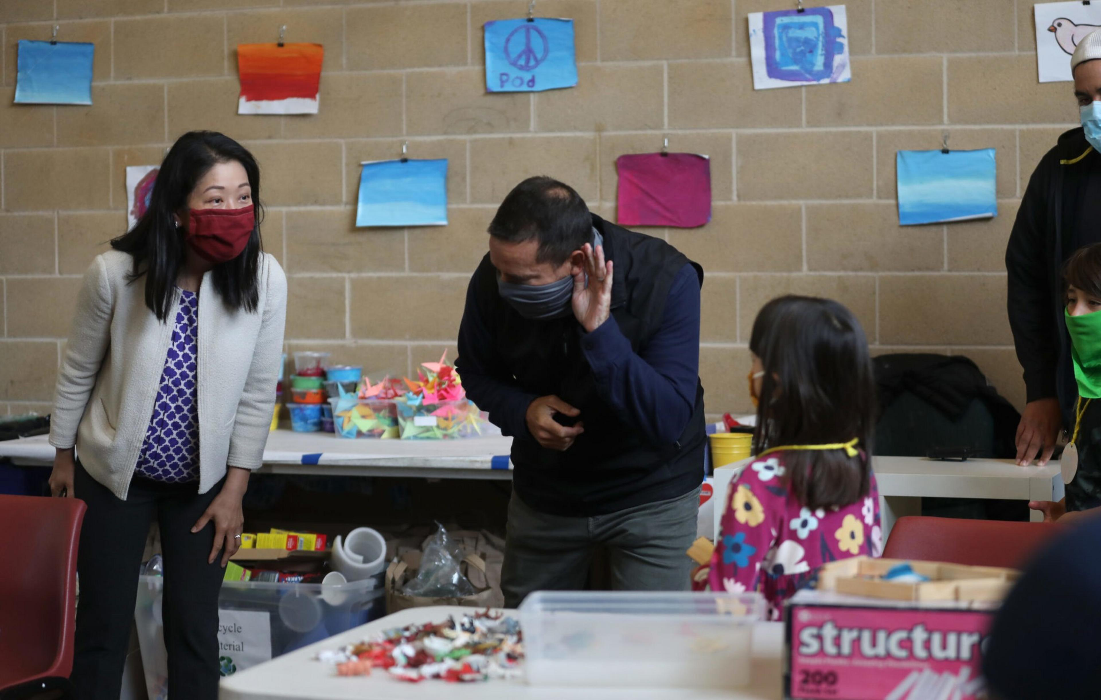 A man and a woman wearing masks interact playfully with children in a classroom decorated with colorful paintings and craft supplies on tables.
