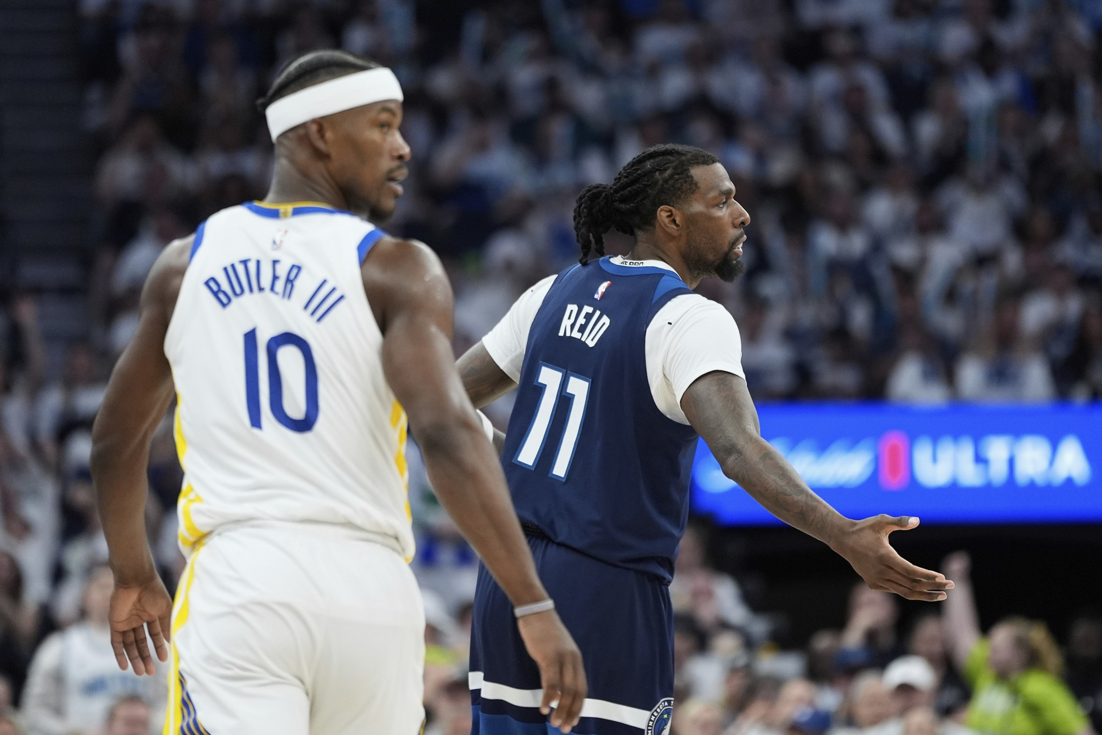 Two basketball players, one in a white jersey (#10) and another in a blue jersey (#11), are in a game setting with a crowd in the background.