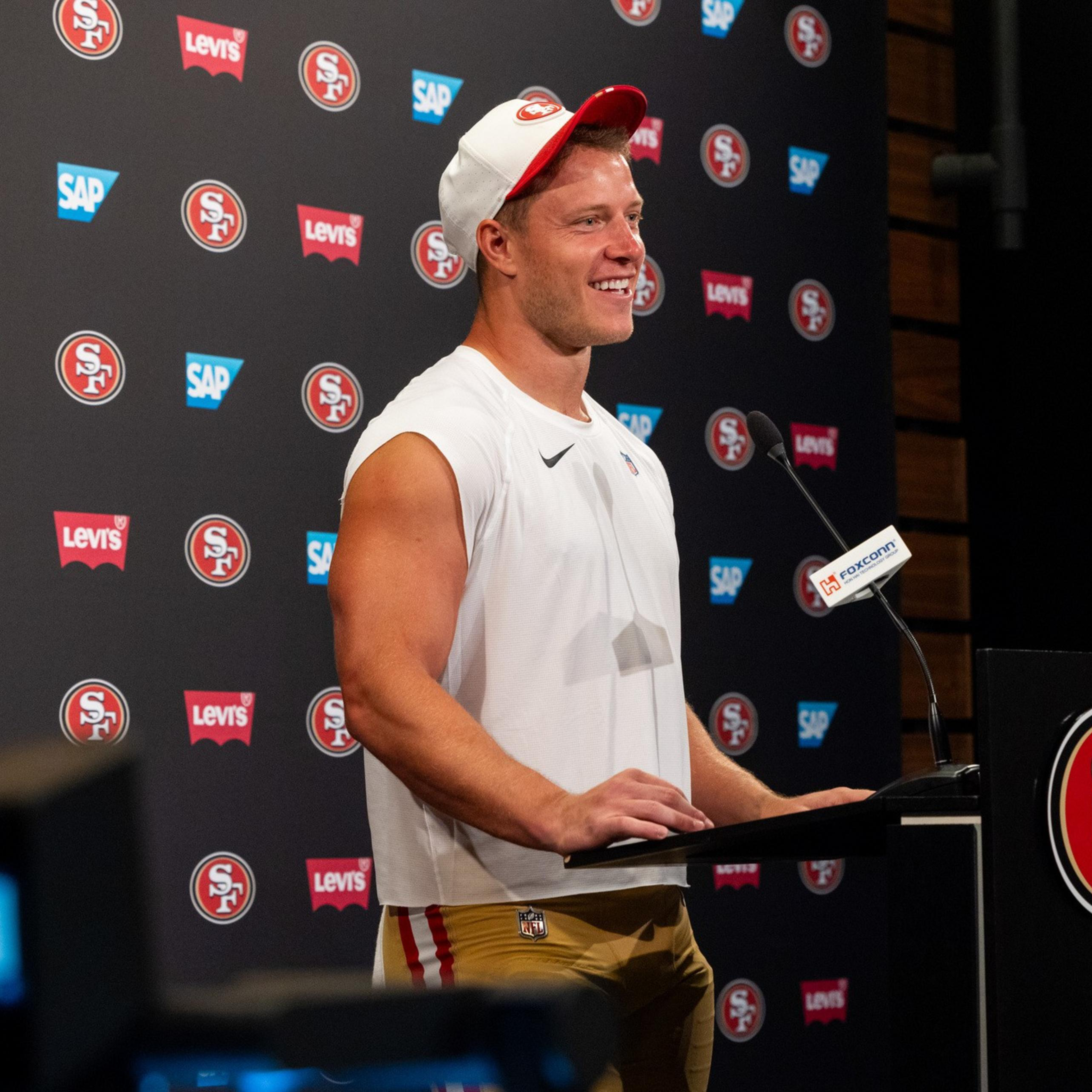 A man in a white sleeveless shirt and beige pants with a white and red cap smiles while speaking at a podium with San Francisco 49ers logos behind him.