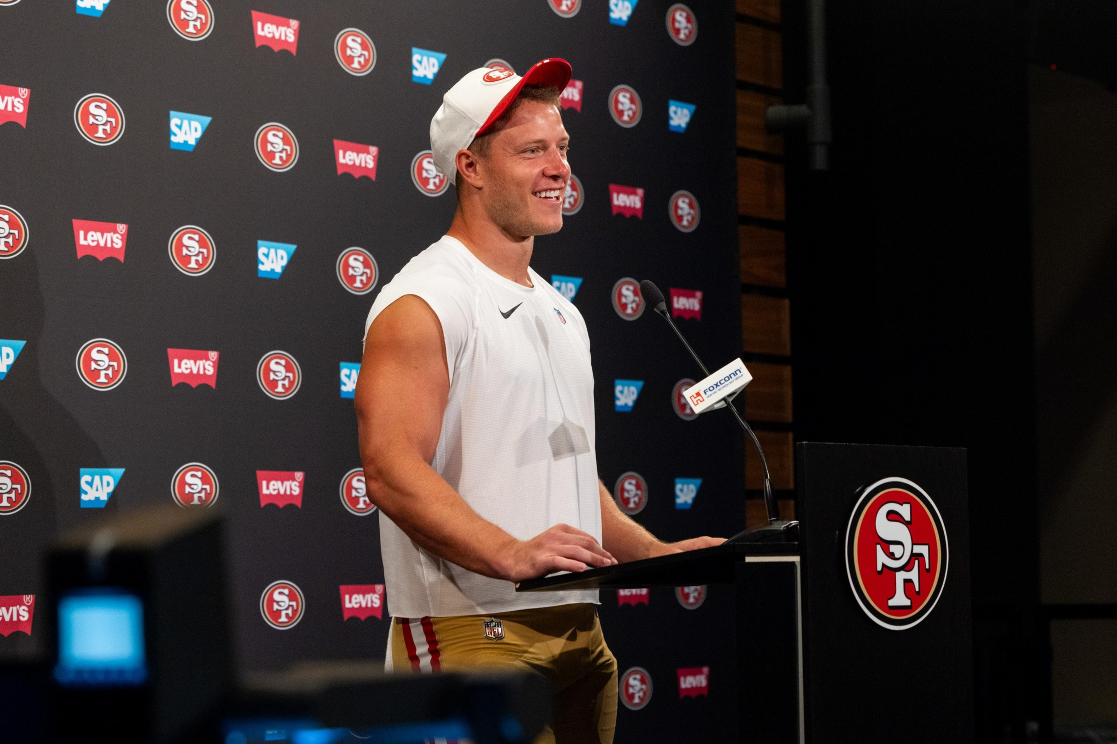 A man in a white sleeveless shirt and beige pants with a white and red cap smiles while speaking at a podium with San Francisco 49ers logos behind him.