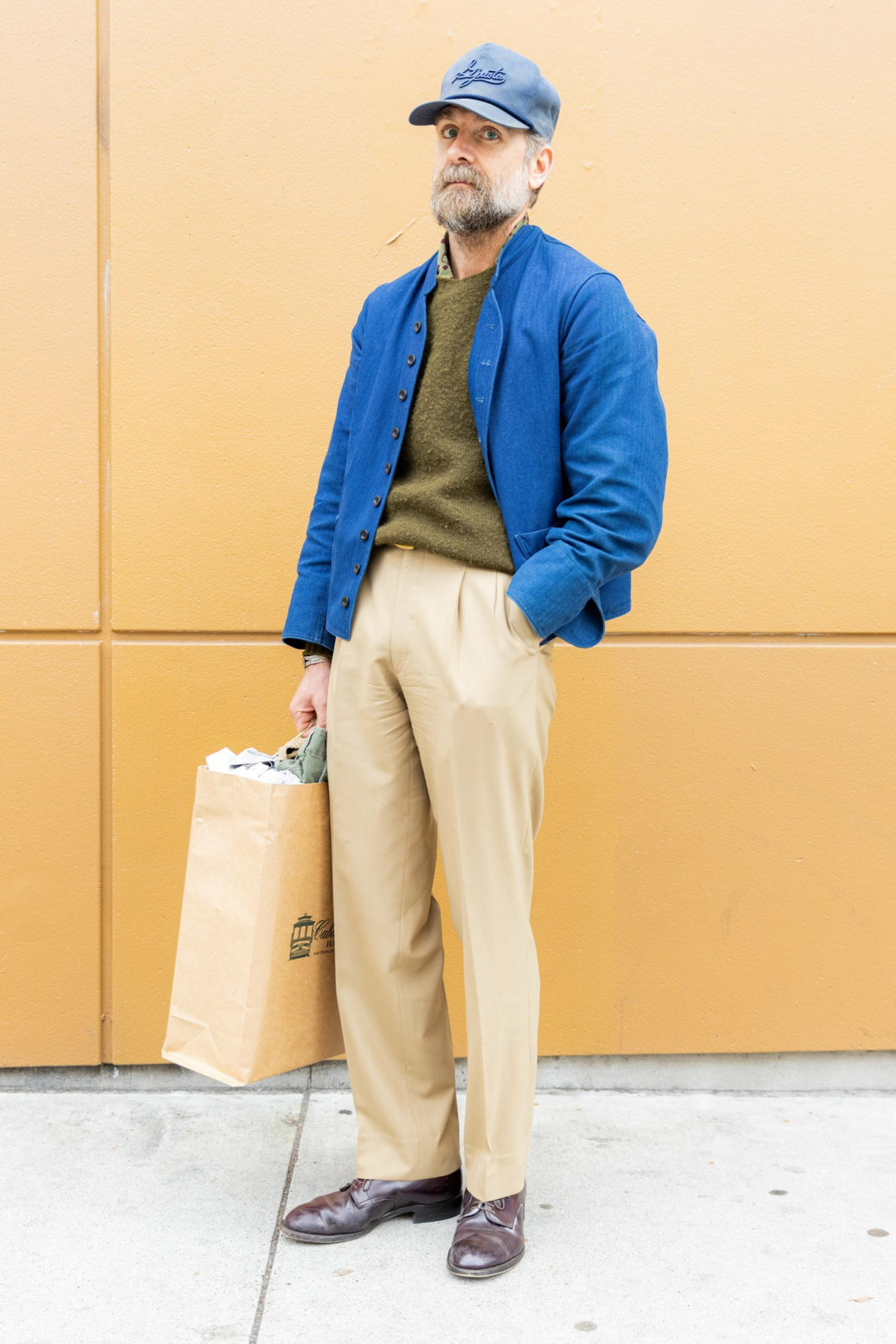 A man wearing a blue jacket, olive green sweater, beige pants, and brown shoes stands against a tan wall holding a brown paper bag in one hand.