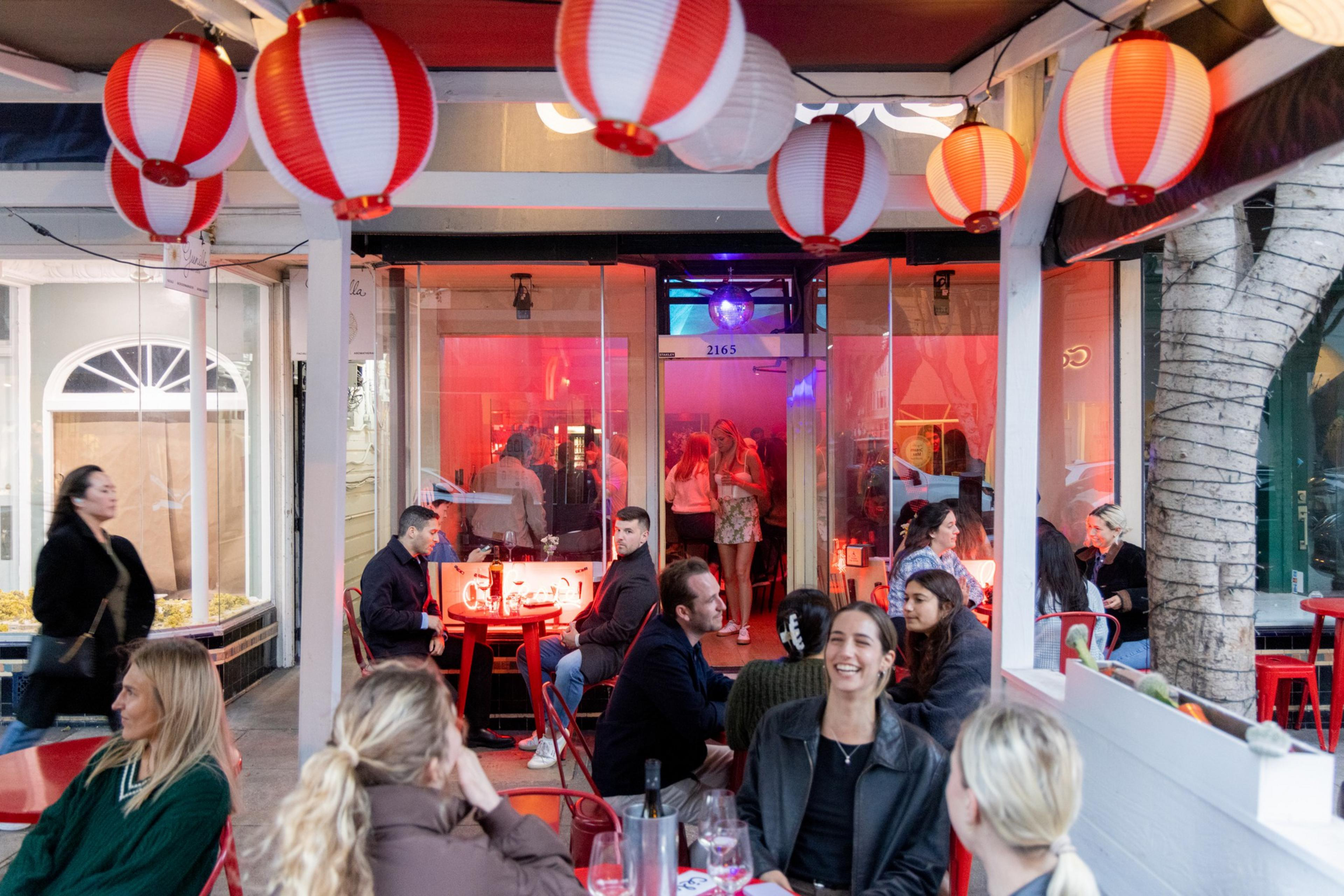 An outdoor restaurant scene with several people seated at red tables, chatting and laughing under red lanterns, while others are walking by and a crowd is inside.