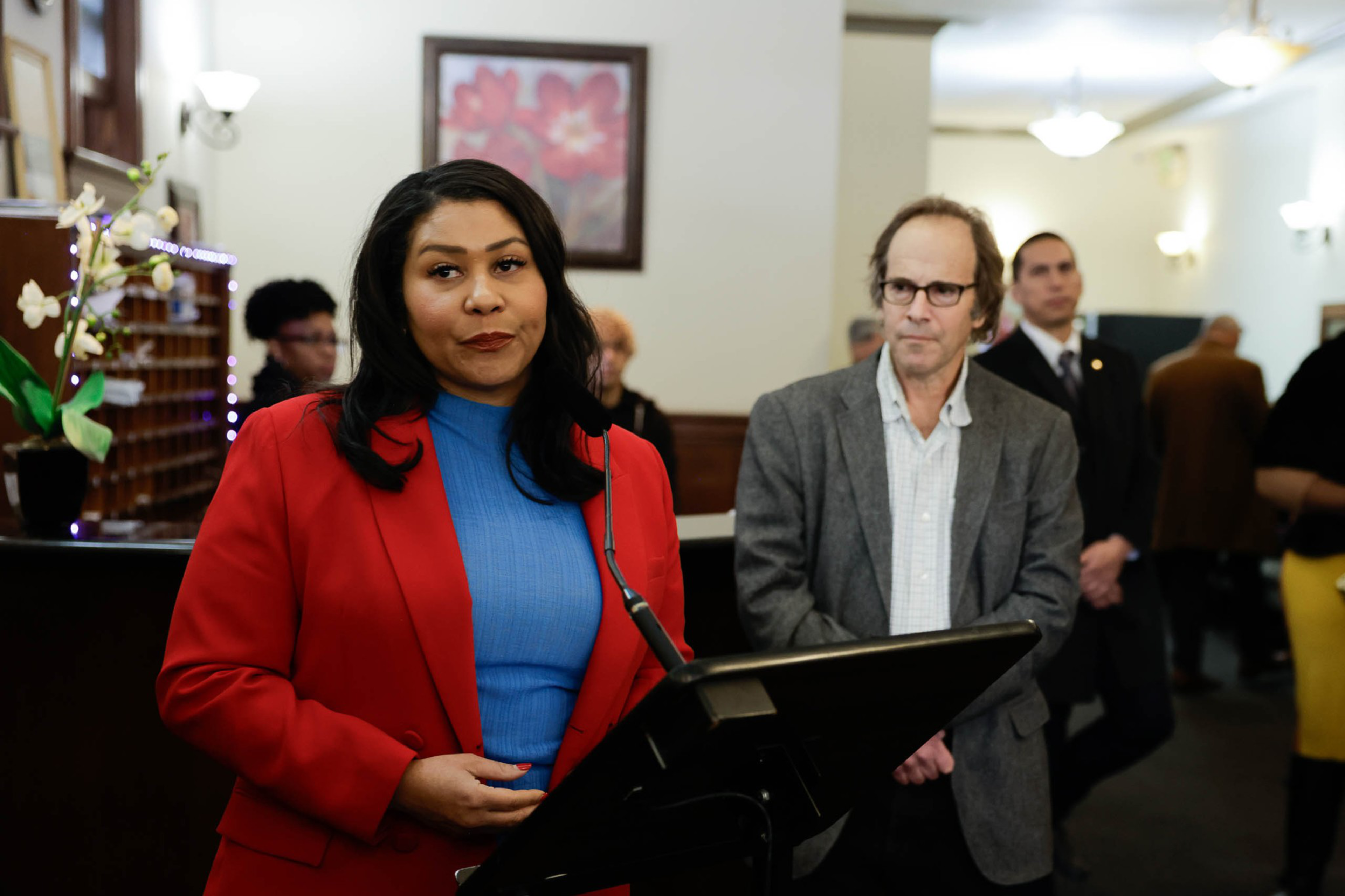 A woman in a red jacket and blue top stands at a podium, flanked by two men in a formal room with onlookers in the background.