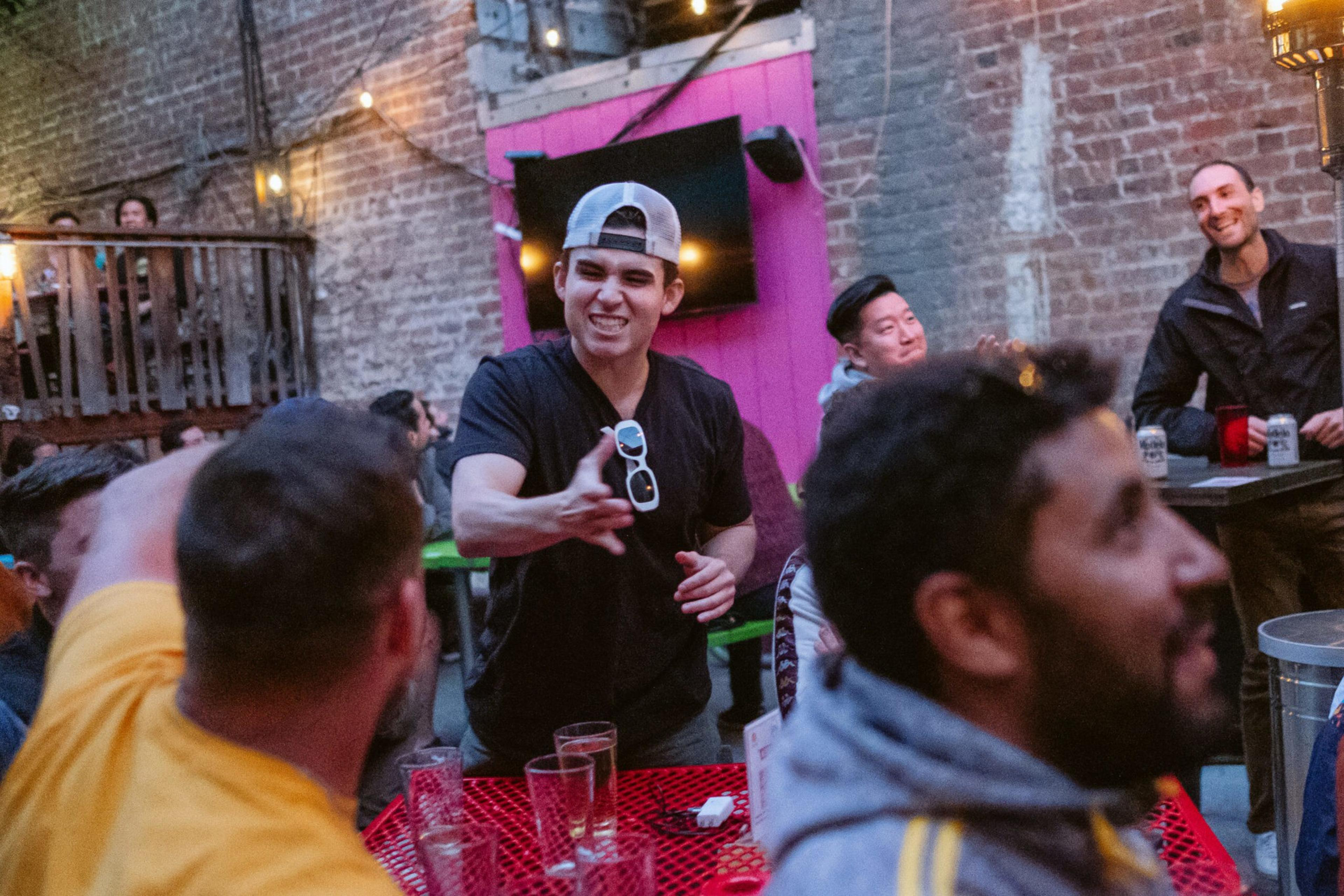 A group of friends socializes outdoors at a table with drinks, one man in a backward cap gestures animatedly, while others smile and chat nearby.