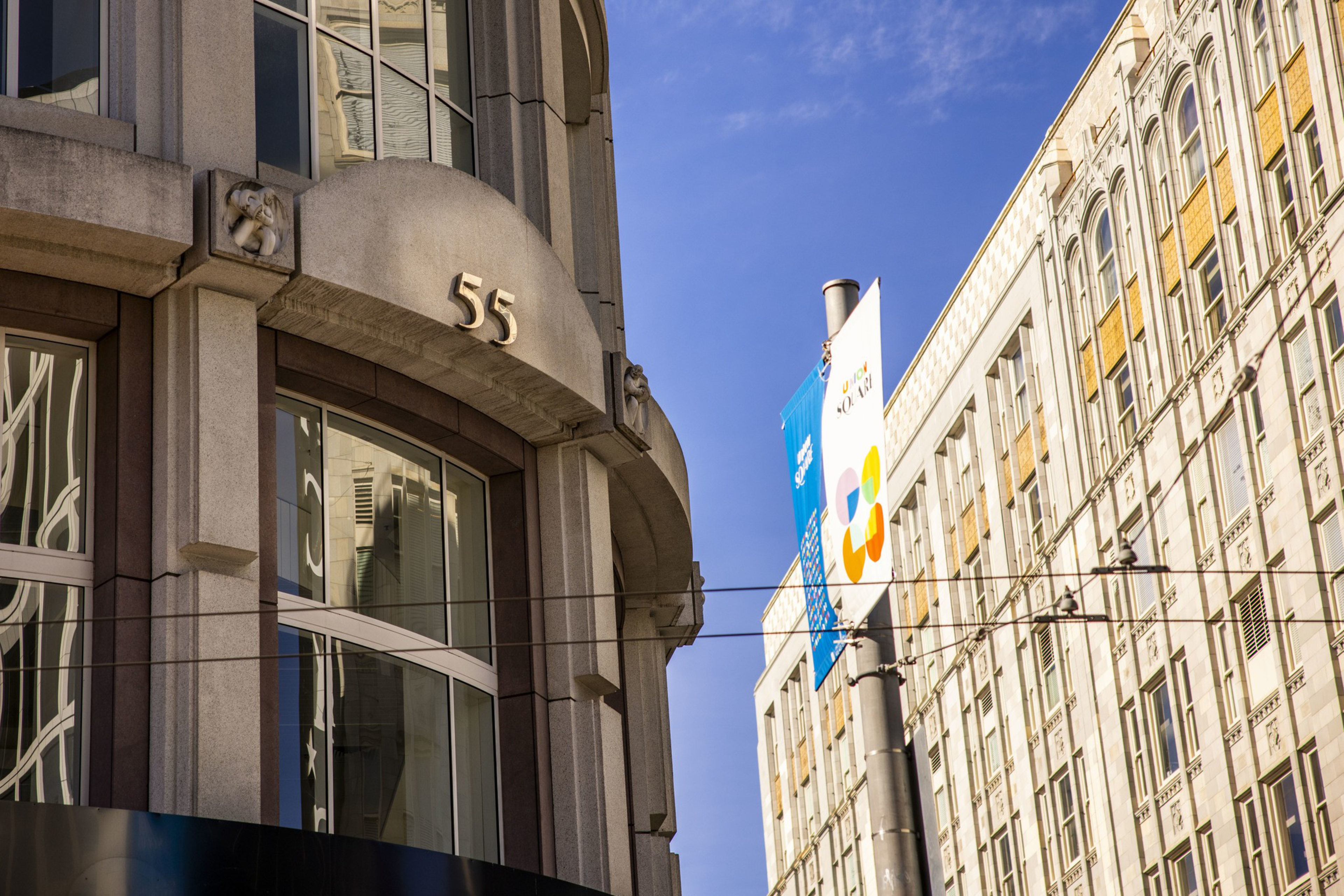 A curved stone building corner displays the number 55 above large windows, with a streetlight and banners against a clear blue sky nearby.