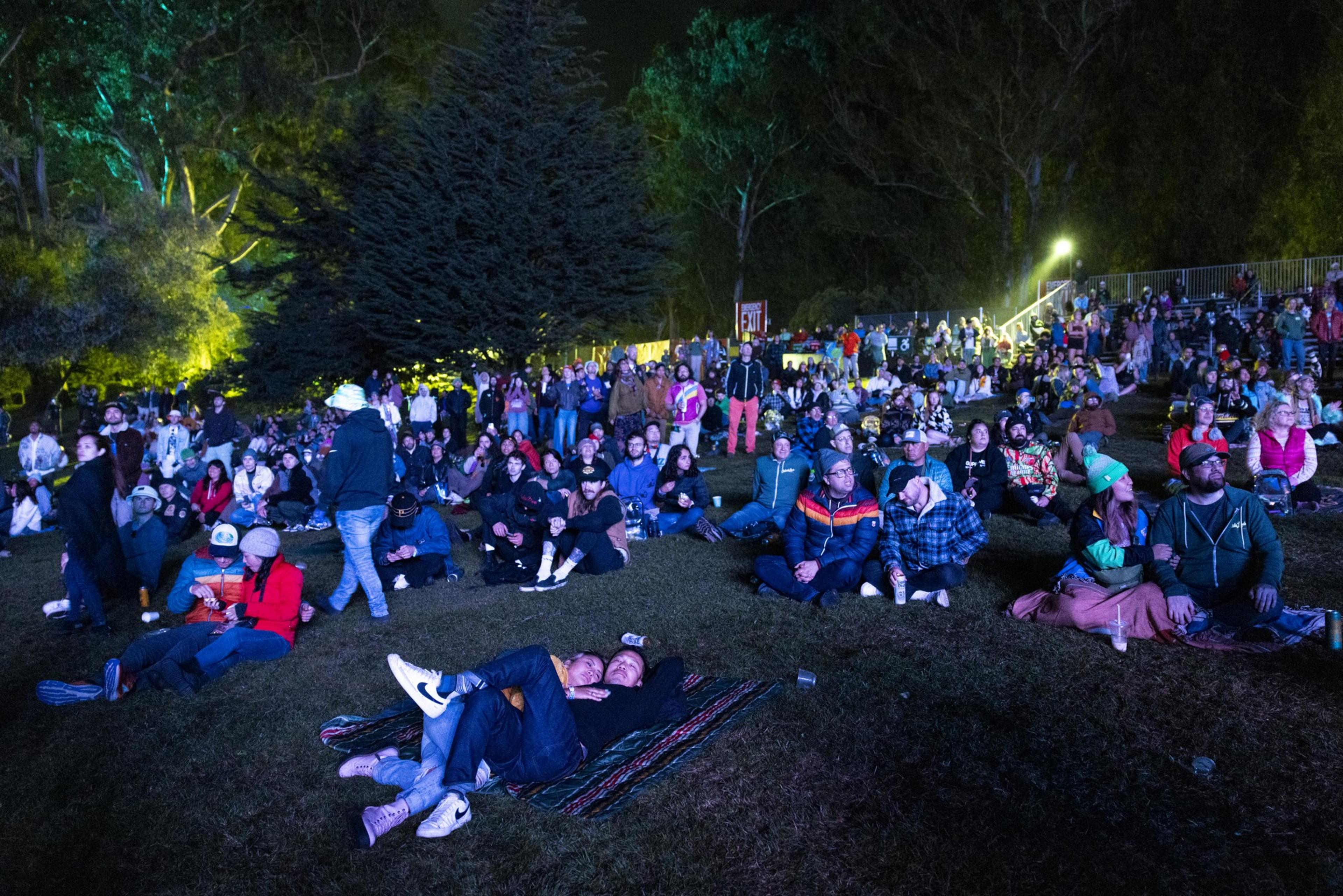 A crowd of people is seated on grass at an outdoor nighttime event, with some lying on blankets and others sitting in groups, surrounded by illuminated trees.