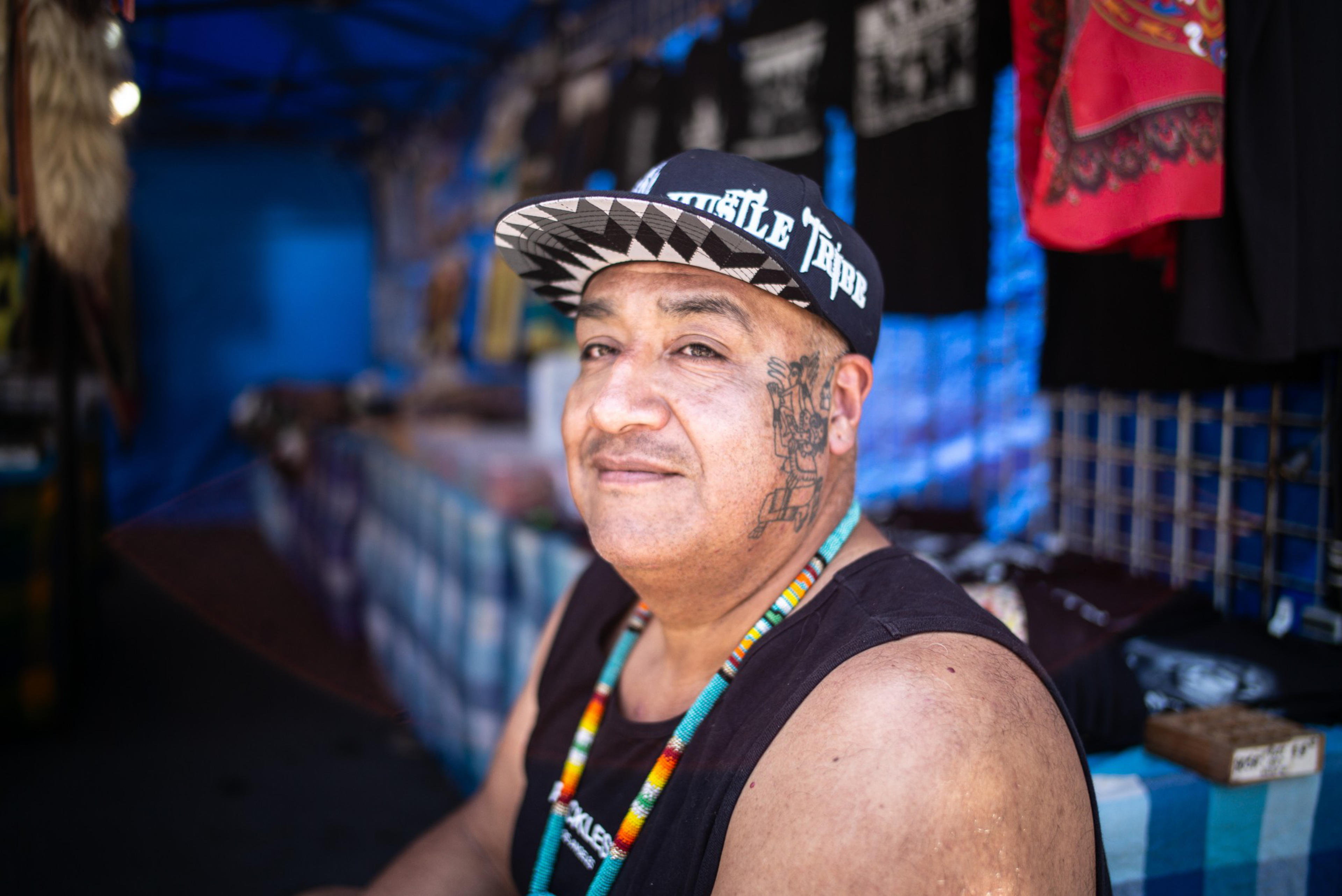 A man with a face tattoo and wearing a patterned cap and beaded necklace sits in front of a booth with colorful fabrics and shirts.