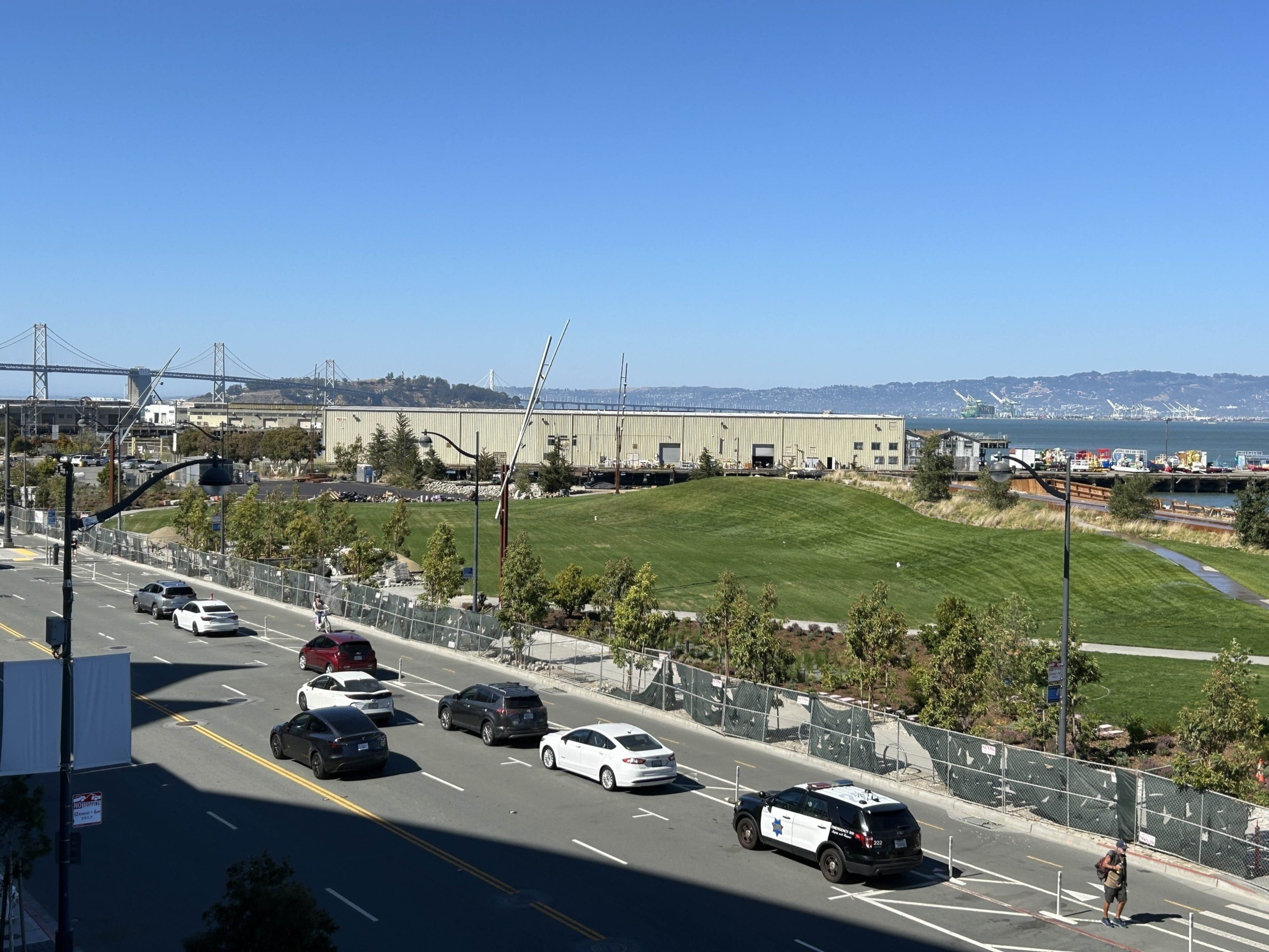 The image shows a street with six cars and a cyclist, a grassy park area, an industrial building, and a distant view of a bridge and cranes against a clear blue sky.