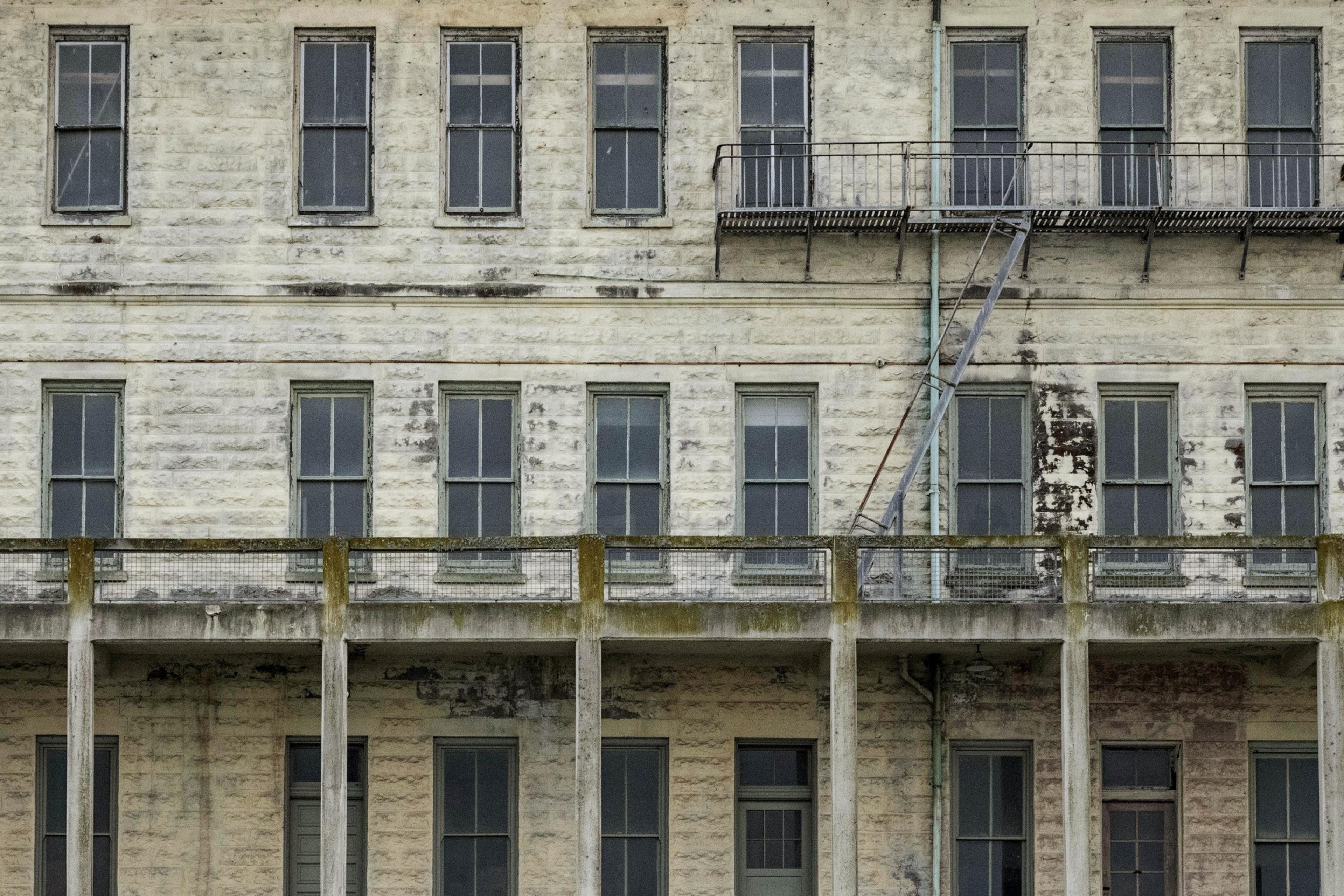 The image shows an aged, weathered building facade with multiple rows of old windows. There's a rusty fire escape stairs and balcony on the upper level.