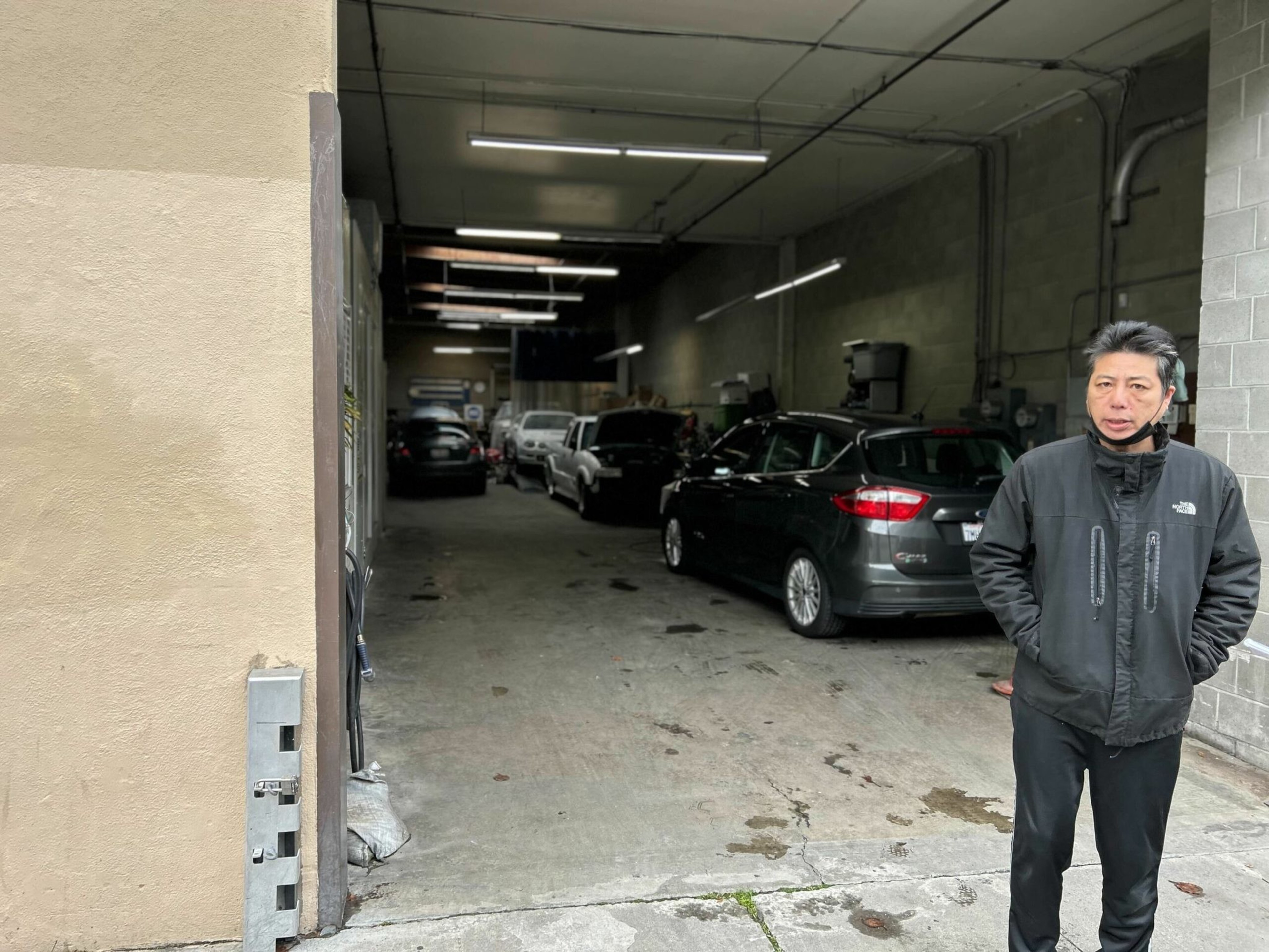 A man in a black jacket stands outside a dimly lit garage with several parked cars inside along a concrete floor and walls.